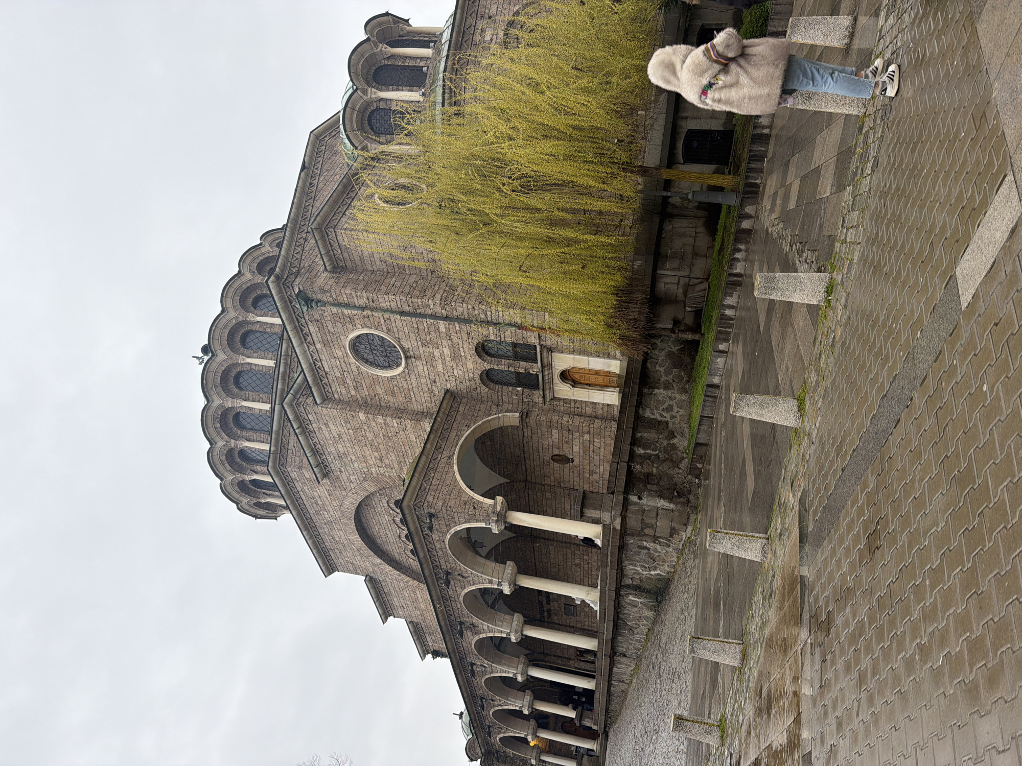 The image depicts the St. Alexander Nevsky Cathedral in Sofia, Bulgaria, on an overcast day. The cathedral's facade, constructed of light-colored stone, features a series of arches supported by columns, as well as rounded windows and domes. A deciduous tree with yellowish-green leaves is positioned in front of the cathedral. A person wearing a light-colored fur coat and blue jeans stands on a cobblestone walkway. Concrete bollards line the walkway.