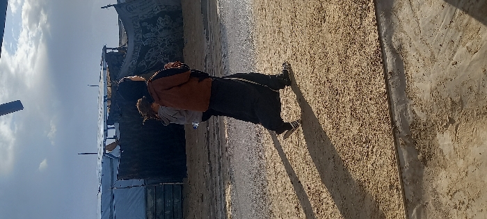The image depicts an outdoor scene in Khan Yunis, Palestinian Territory, under a partly cloudy sky. Two individuals walk from left to right across a light brown, sandy ground with visible wet patches and shallow puddles reflecting light. The person in the foreground wears a brown jacket, dark trousers with a white stripe, and black-and-white striped athletic shoes, casting a long shadow to their right. Behind them, a second individual wears a grey hooded sweatshirt and carries a clear plastic bottle in their right hand. In the background, a series of rudimentary structures constructed from materials such as corrugated metal sheets, tarpaulins, wood, and patterned fabric are visible. A dark, rectangular object is partially cropped at the top center of the frame.