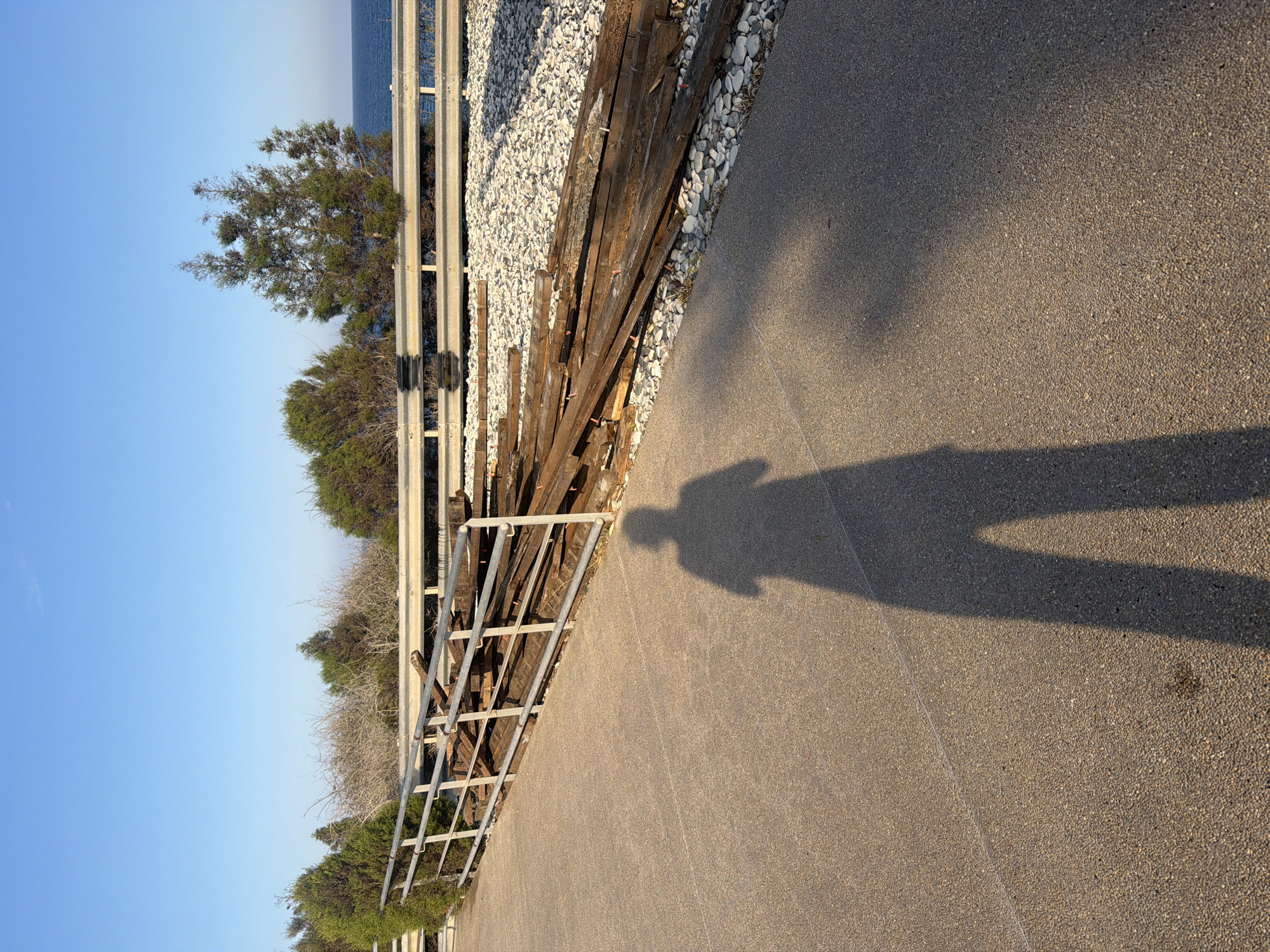 In Pervolia, Cyprus, a shadow of a person with legs and torso is cast on a textured, light brown paved surface. To the left and extending into the background, a metal railing with horizontal bars follows an upward slope. Beyond the railing, a pile of weathered wooden planks lies on a bed of light-colored gravel. In the background, green bushes and trees are visible, with a clear blue sky above. In the far distance, the blue sea meets the horizon.
