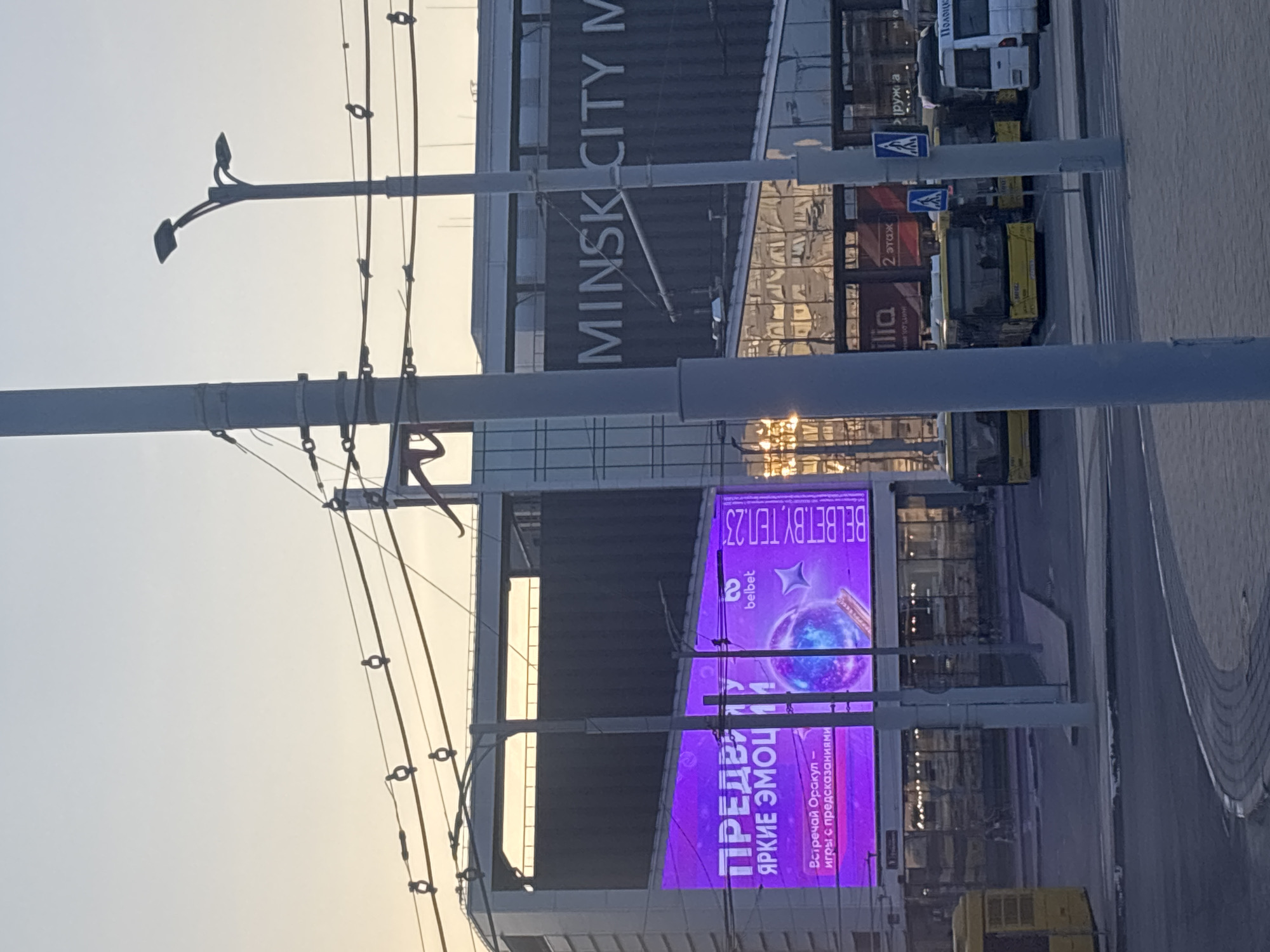 The image shows a wide shot of a city street in Minsk, Belarus, likely near Minsk City Mall, as evidenced by the prominent "MINSK CITY M" signage. The scene is dominated by a large digital billboard displaying a vibrant purple advertisement for "belbet," featuring text in Russian, including "ПРЕДВЫШУ ЯРКИЕ ЭМОЦИИ" (Experience Bright Emotions) and "Встречай Оракул - игры с предсказаниями" (Meet the Oracle - games with predictions). The billboard also shows an image of a glowing orb and a star.
In the foreground, several utility poles and overhead wires crisscross the frame. Behind the billboard, a modern building with glass facades reflects the sky and surrounding structures. Several yellow buses are parked in what appears to be a bus stop or station area, with passengers visible through their windows. A white van is also parked nearby. The sky suggests it is either early morning or late evening due to the soft, diffused light. The overall atmosphere is urban and bustling, capturing a moment in the daily life of the city.