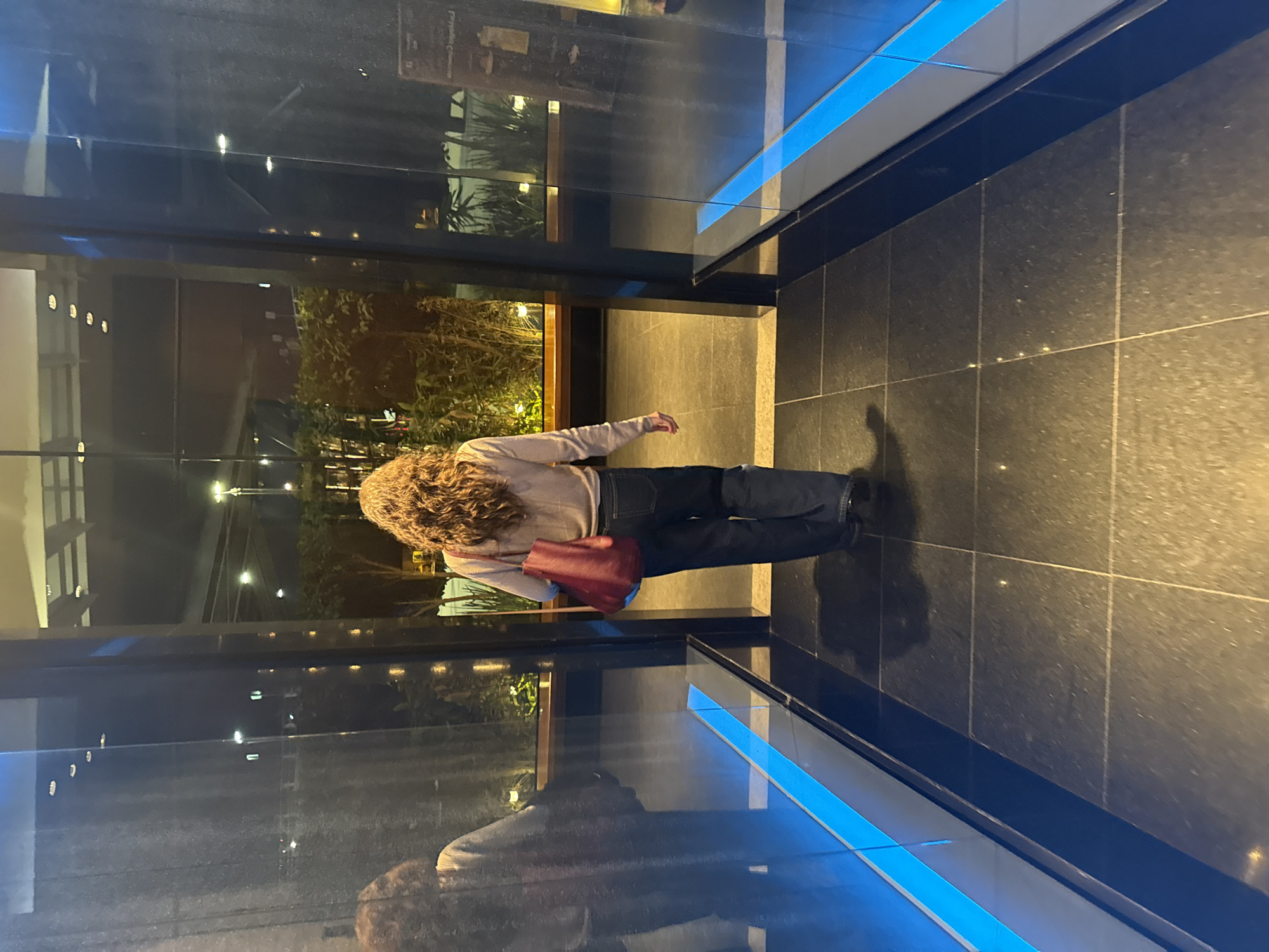 A woman with curly hair, wearing a light-colored sweater, blue jeans, and black shoes, stands on a shiny black tile floor within a modern building in Puebla, Mexico. She has a red purse slung over her shoulder. The building features glass walls that reflect outdoor vegetation and streetlights, creating a mirrored effect. Blue lighting accents the lower part of the glass wall. The overall ambiance suggests an upscale or contemporary commercial space.