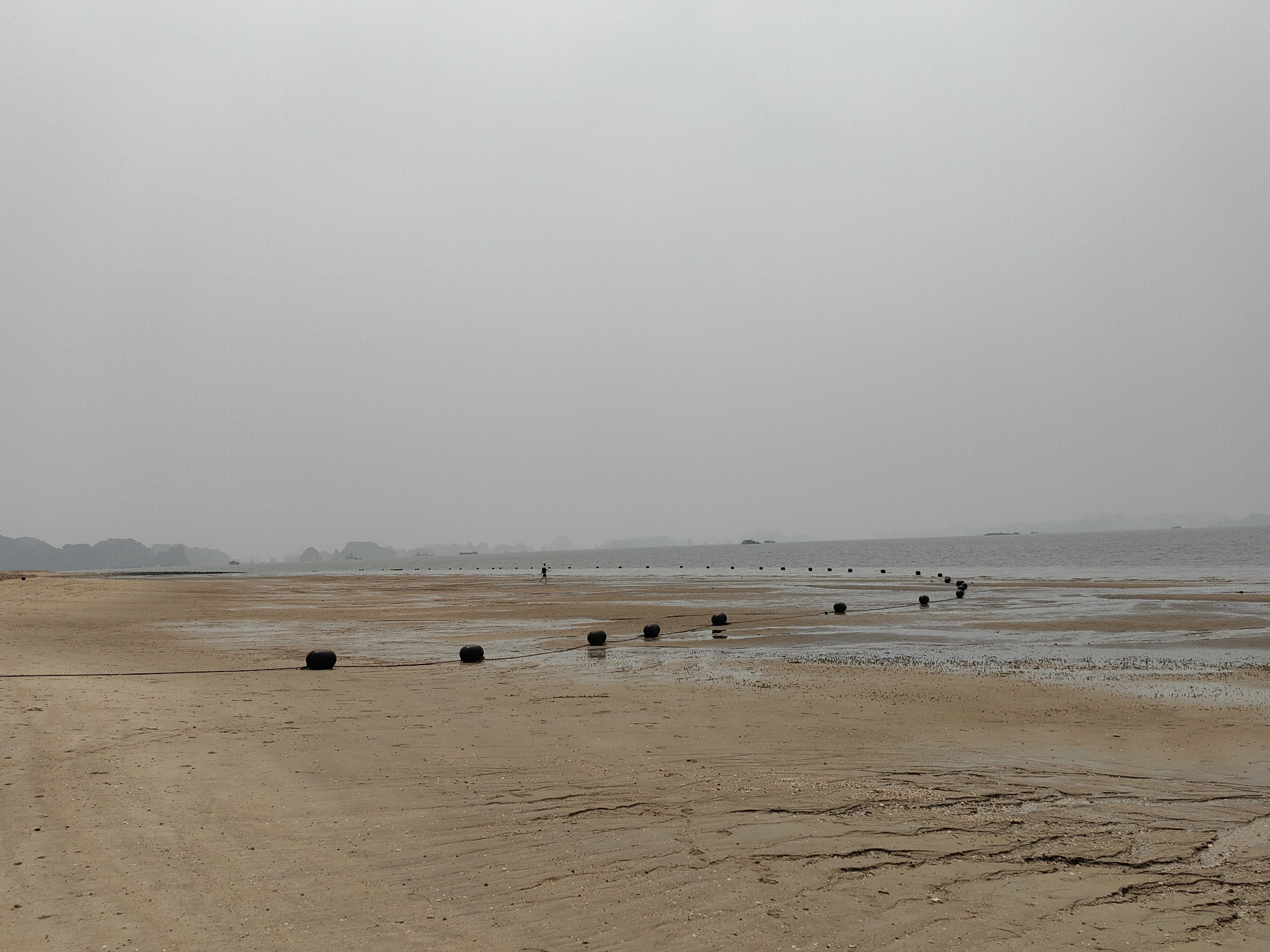 The image shows a beach at Bai Chay Ward, Vietnam. The beach is wide and sandy with tidal patterns visible in the foreground. A series of dark, spherical buoys are strung together in a line, extending from the left foreground into the water towards the right. The water appears calm, with a hazy sky overhead. In the distance, there are blurred outlines of hills or islands. A single person is visible in the distance, walking along the beach towards the water.