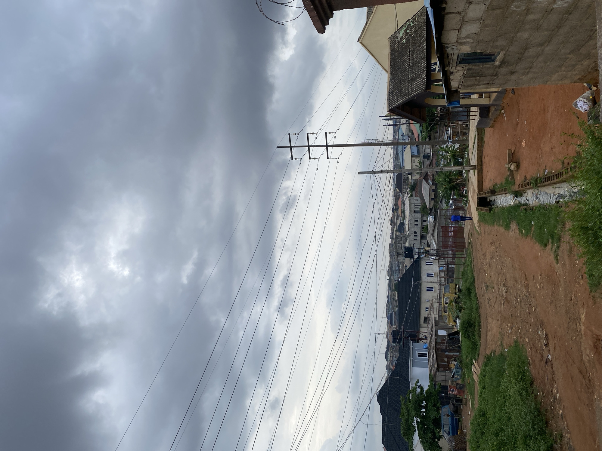 The image depicts a street scene in Lagos, Nigeria, under a heavily overcast sky. Power lines crisscross the sky above a densely built urban landscape. Buildings of varying heights and construction materials line the street. A dirt path with patches of grass runs along the right side of the frame. One person is visible in the distance, walking along the street. Debris is visible along the side of the road, indicative of a developing urban environment.