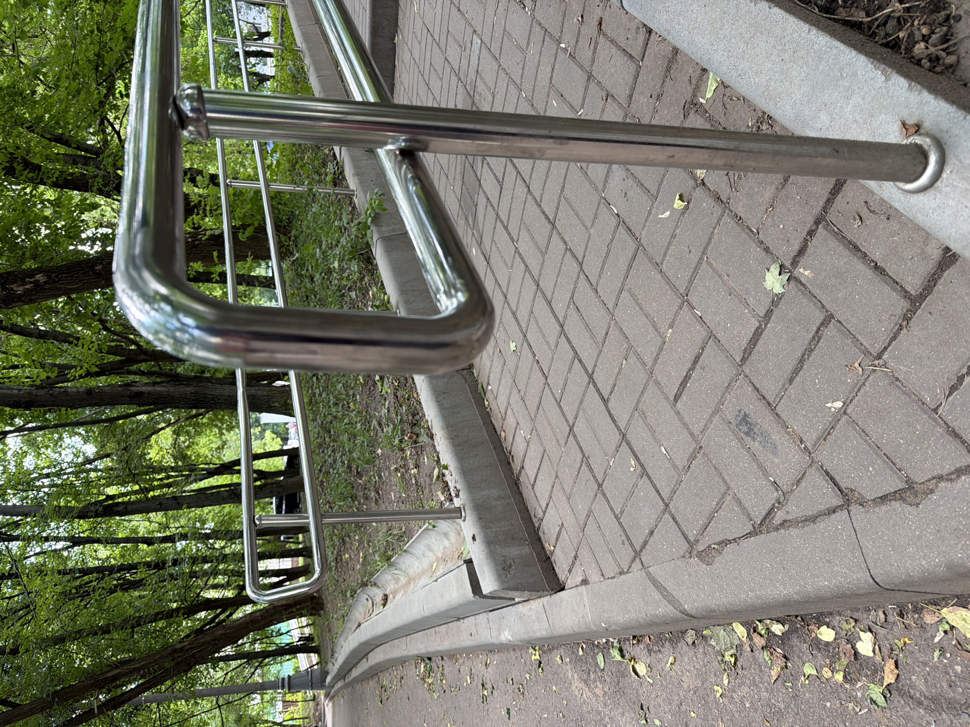The image shows a paved walkway with a metal handrail in a park-like setting. The walkway is made of rectangular pavers laid in a herringbone pattern. A concrete curb runs alongside the walkway, separating it from a patch of grass and trees. Fallen leaves are scattered on the ground. The handrail is made of polished metal tubes and is supported by vertical posts. In the background, trees with green foliage are visible, suggesting a warm season. The scene appears to be a public pathway, possibly in a park or pedestrian area. While no specific landmarks are visible to pinpoint the exact location within Moscow, the presence of mature trees and paved pathways is common in Moscow's many parks and green spaces, such as Gorky Park or Sokolniki Park.
