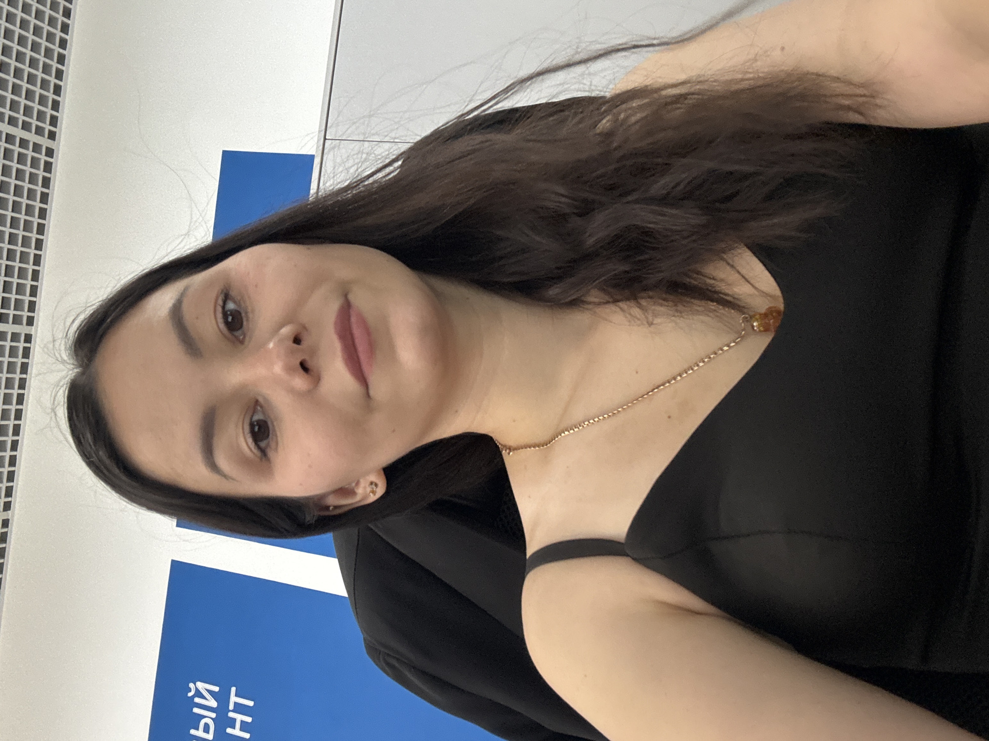 A medium shot shows a young woman with long, dark brown hair sitting in an office environment. She is wearing a black sleeveless top and a gold necklace with a small amber pendant. A black blazer is visible behind her. The background includes a blue and white sign with Russian text and a white wall with a grid pattern on the ceiling. The image is taken from a slightly low angle, looking up at the woman. The location is identified as Vavilovo, Russia.