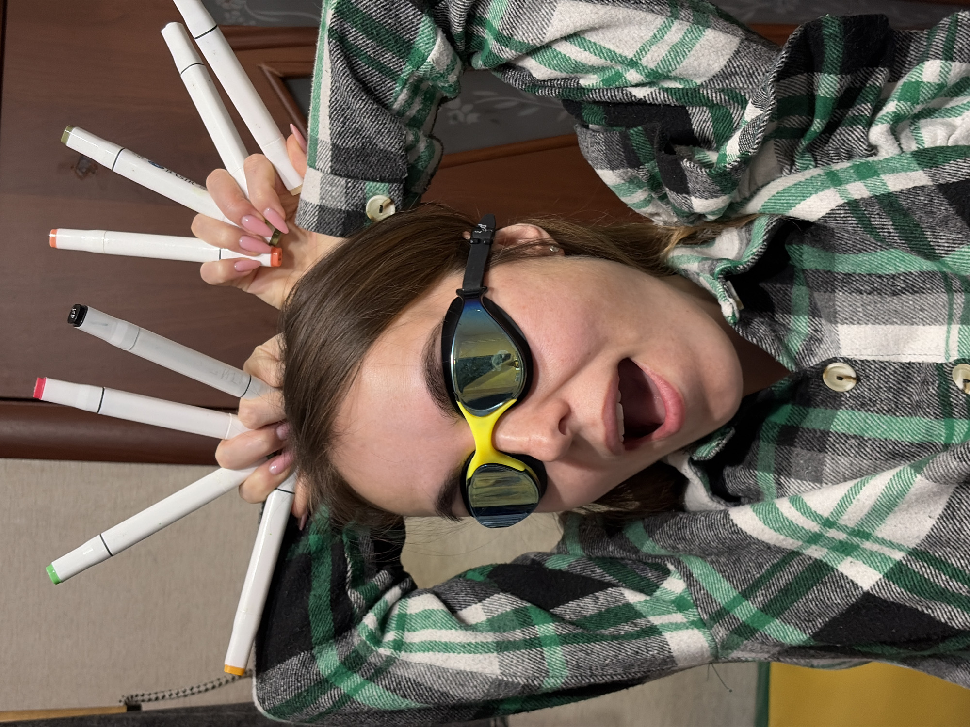 A woman wearing swimming goggles on her forehead and a green plaid shirt is holding several white markers in her hands. The markers have different colored caps. She is looking at the camera with her mouth open. The location is not discernible as Izhevsk, Russia, from the image alone.
