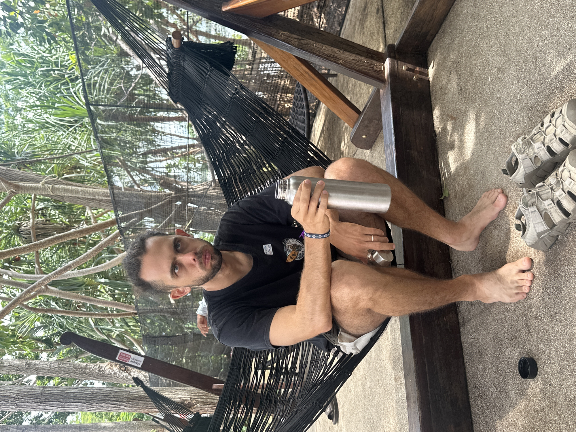 A man is sitting in a black hammock outdoors, holding a silver metal water bottle. He is wearing a black t-shirt and light-colored shorts. His feet are bare, and a pair of grey sandals are visible on the ground next to him. The hammock is suspended from a wooden structure, and behind the man, palm trees and foliage are visible, suggesting a tropical setting. The ground appears to be covered in a textured material, possibly concrete or gravel. The lighting suggests it is daytime, and the weather appears to be clear and sunny. The man is looking towards the camera with a neutral expression. A small white sticker with red text is visible on the wooden support beam of the hammock. The text on the sticker appears to be in Thai and English, and may indicate a weight limit or safety instruction, though it is difficult to read clearly. The overall scene depicts a man relaxing in a hammock in a tropical environment.
