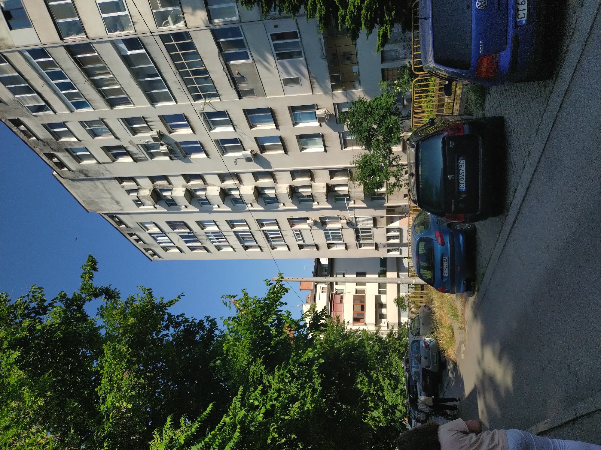 The image depicts a residential street in Stara Zagora, Bulgaria, identifiable by "CT" prefixes on multiple vehicle license plates. The view is from a pedestrian's perspective, looking down a street lined with parked cars, apartment buildings, and trees under a clear blue sky.
In the left foreground, a person is partially visible from the waist down, wearing a light-colored top and white pants, walking away from the camera on a paved sidewalk. Further ahead on the sidewalk, two to three additional individuals are walking, with one appearing to push a stroller, partially obscured by parked vehicles.
A large, dense deciduous tree with vibrant green foliage occupies the left side of the frame, casting shadows onto the street and sidewalk. Behind this tree, smaller, lighter-colored residential buildings with reddish-brown roofs are visible in the background.
The right side of the image features a tall, multi-story apartment building with a light grey, weathered facade. The building has numerous windows, some small open balconies, some enclosed glass balconies, and multiple external air conditioning units. Below the building, a yellow metal fence separates the sidewalk from an overgrown area with small evergreen trees and dry grass.
Several vehicles are parked along both sides of the street. On the right, parked away from the camera, are a dark blue Volkswagen Golf (partially visible with a Bulgarian "CT" plate) and a black Peugeot 307 SW station wagon with license plate CT 0657 BC. On the left side of the street, parked in a row, are a blue Peugeot 206 hatchback (license plate CT 7986 BA), a silver hatchback (partially visible license plate CT 0492), and another silver car with a Sofia-registered license plate CA 4991 PP.
The sky is a clear, bright blue, with a small, dark object (likely a bird or insect) visible in flight in the upper right. Strong sunlight illuminates the scene, creating distinct shadows across the street and on the buildings, indicating daytime conditions. A light-colored utility pole with overhead lines is present in the mid-ground on the left.