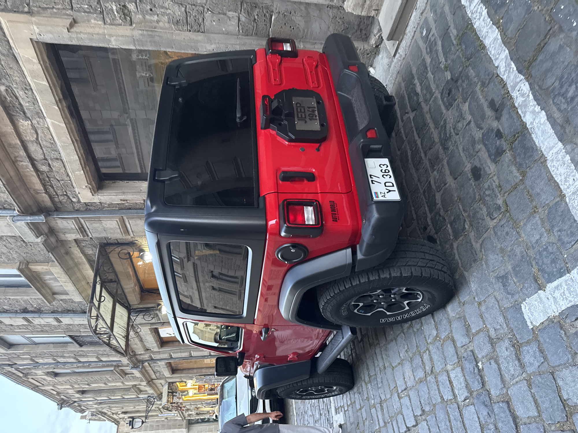 A bright red Jeep Wrangler two-door is parked on a cobblestone street in the city of Sabail Raion, Azerbaijan. The Jeep is facing away from the viewer, and its rear is prominently displayed. It has a black hardtop, black fender flares, and black side steps. The Jeep's license plate is visible and reads "77 YD 363 AZ". A decorative plate with "JEEP 1941" is mounted on the spare tire cover. To the left of the Jeep, a silver Kia SUV is partially visible, with a person in a grey shirt and white pants standing next to it. The background consists of a stone building with arched windows and awnings, typical of the architecture in the Old City of Baku, which is part of Sabail Raion. The lighting suggests it is daytime, possibly late afternoon or early evening.