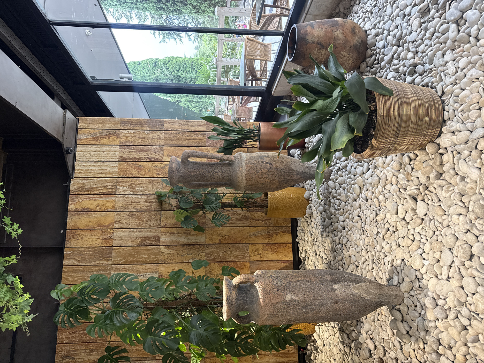 The image depicts an interior space with an outdoor view through large glass windows. The interior features a stone wall with vertical wooden paneling, a floor covered in white pebbles, and several large, antique-style ceramic vases. Plants are strategically placed throughout the space, including a large Monstera plant. In the foreground, a rattan planter holds a lush green plant. The outdoor view reveals a patio area with wicker furniture and tables, surrounded by cypress trees. The overall aesthetic is rustic and natural, suggesting a restaurant or a well-decorated indoor garden area. The location is identified as Yalta, Ukraine.