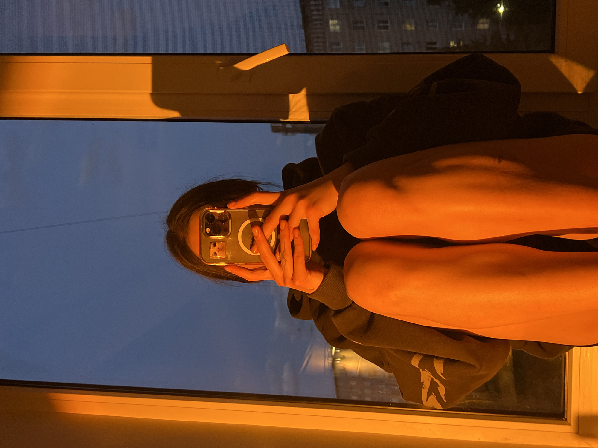 A person sits in a window seat, legs drawn up, facing a window with a view that shows a building across the street. The person is wearing a hooded sweatshirt and is holding a phone to take a picture. The lighting appears to be a combination of natural light from the window and warm artificial light. The city visible through the window is Ternopil, Ukraine.