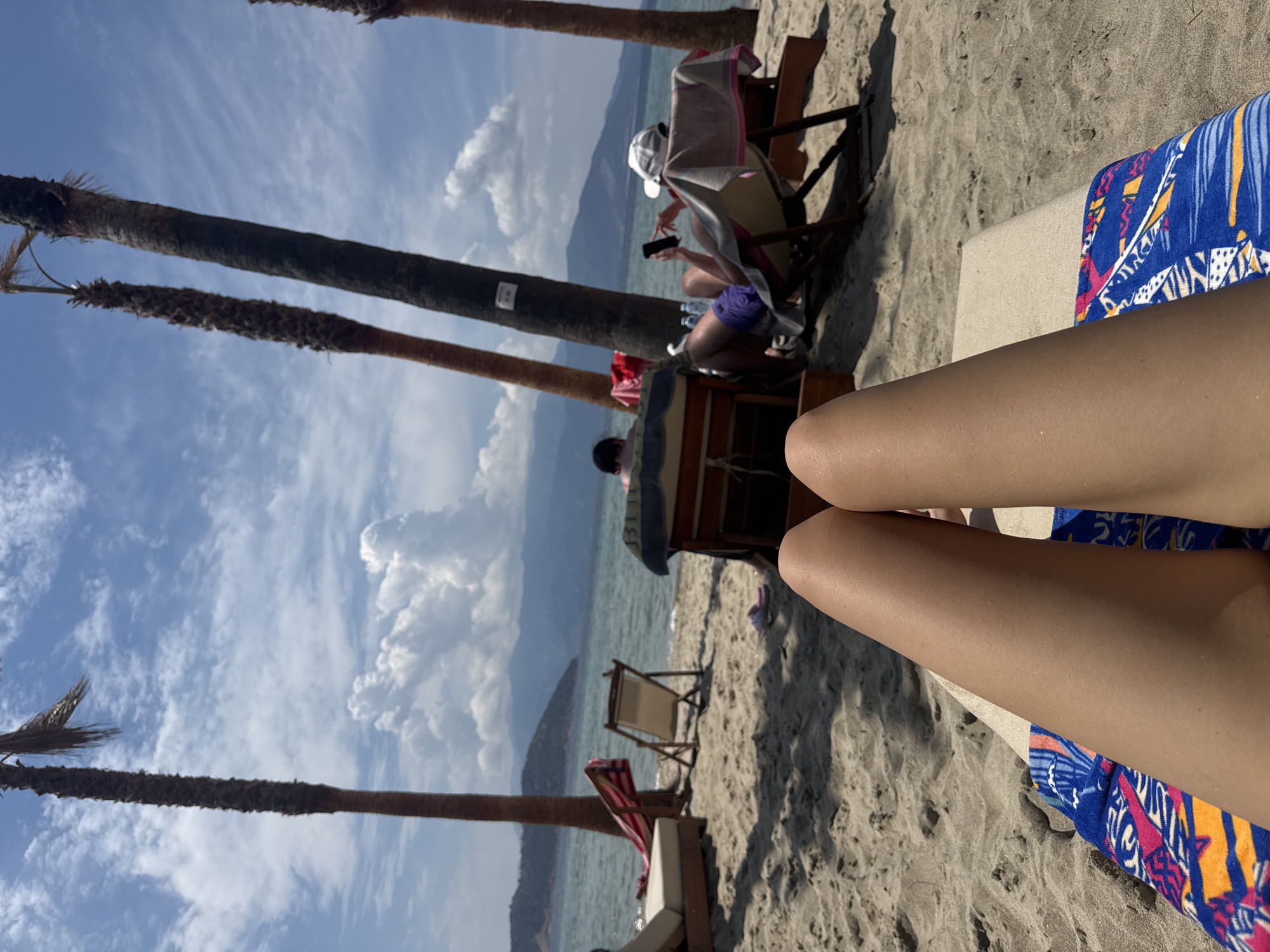 The image shows a first-person perspective of someone relaxing on a sun lounger on a sandy beach in Keramoti, Greece. In the foreground, the legs of the person are visible, resting on a colorful towel. The background features a tranquil sea, distant mountains, and a bright sky with scattered clouds. Several palm trees are strategically placed along the beach, providing shade. Other beachgoers can be seen in the middle ground, seated on loungers and enjoying the seaside atmosphere. One person is looking at their phone, while another appears to be resting. The overall scene depicts a serene and idyllic beach setting.
