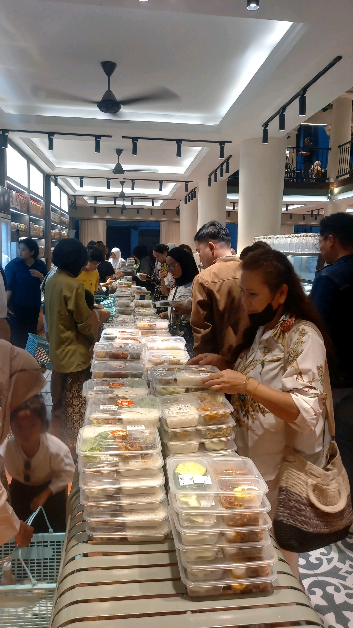 The image depicts a crowded indoor setting, likely a food catering service or a buffet-style restaurant in Medan, Indonesia. Several people are standing around tables laden with stacked transparent food containers. The containers hold various food items, including rice and side dishes, suggesting a meal service. Some individuals are reaching for or holding containers, indicating they are selecting or purchasing food. The environment features modern interior design with white ceilings, track lighting, and a visible air conditioning unit. Columns and a balcony area are also visible in the background, suggesting a multi-level establishment. The overall scene conveys a busy and active food distribution or consumption event.