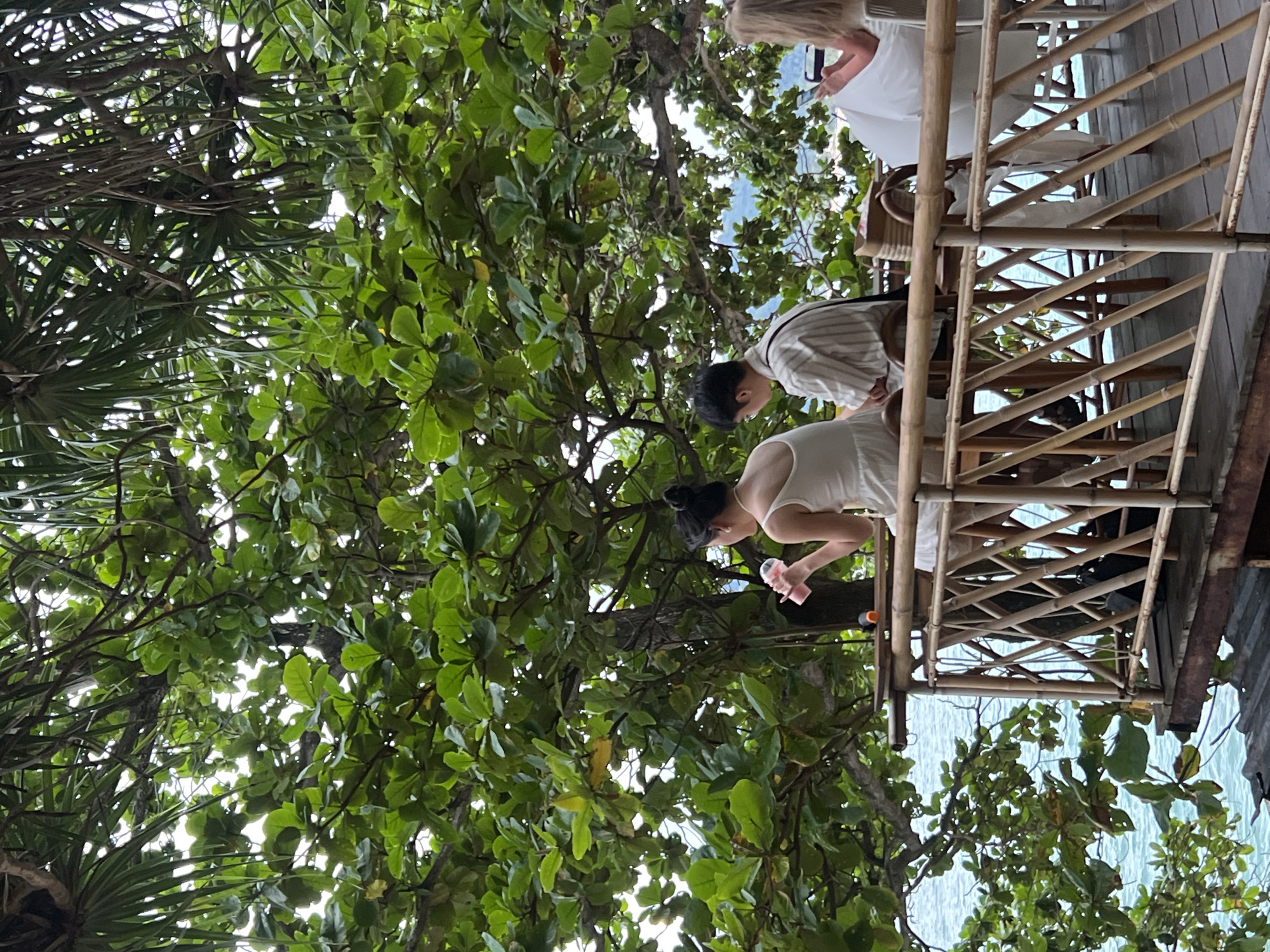 In Karon, Thailand, two individuals are seated on a bamboo balcony overlooking the ocean. A woman in a white dress is on the left, holding a pink cup. Next to her is a man in a striped shirt. To the right, a person in a white outfit is visible, looking at a phone. Lush green foliage partially obscures the view, suggesting a tropical setting. The water is a clear blue, and distant hills are visible. The balcony is constructed of bamboo and overlooks a wooden platform.