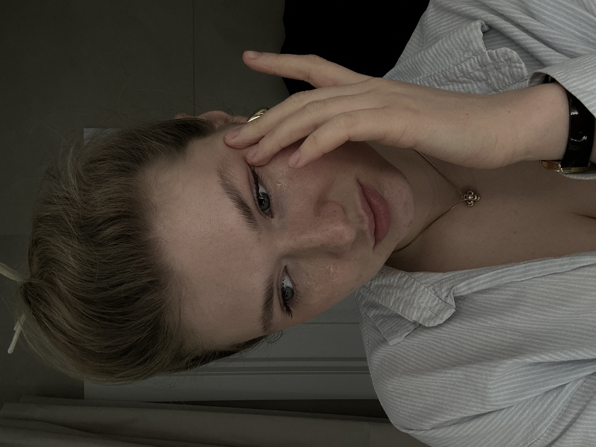 A close-up, low-angle shot depicts a woman with light brown hair partially obscuring her face with her hand. She has fair skin, light-colored eyes, and is wearing a light blue striped shirt. A gold ring is visible on one of her fingers. A gold necklace with a clover-shaped pendant is around her neck, and a black watch is on her wrist. The background is dark, making it hard to see environmental details, but a white object can be seen behind her head. There is no indication of the location being Kazan, Russia.