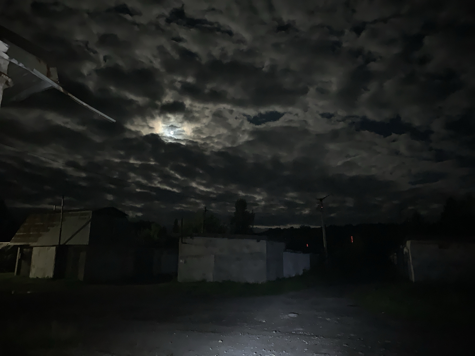 The image captures a nighttime scene with a partially visible moon peeking through dark, cloudy skies. In the foreground, a rough, unpaved surface resembling a road or lot is illuminated by a bright light source, possibly a flashlight or camera flash, from the lower left corner. To the left, there is a structure with a corrugated metal roof and a lighter-colored, possibly concrete or cinder block, wall. Further to the right, another, darker structure is visible, with hints of grass or vegetation growing around its base. In the middle ground, several low, boxy structures that appear to be garages or sheds are dimly lit. A utility pole with what looks like a small, red light is visible to the right of center. The background consists of dark trees and the night sky. The overall impression is of a somewhat dilapidated, possibly industrial or rural, area at night. The location is identified as Asha, Russia.