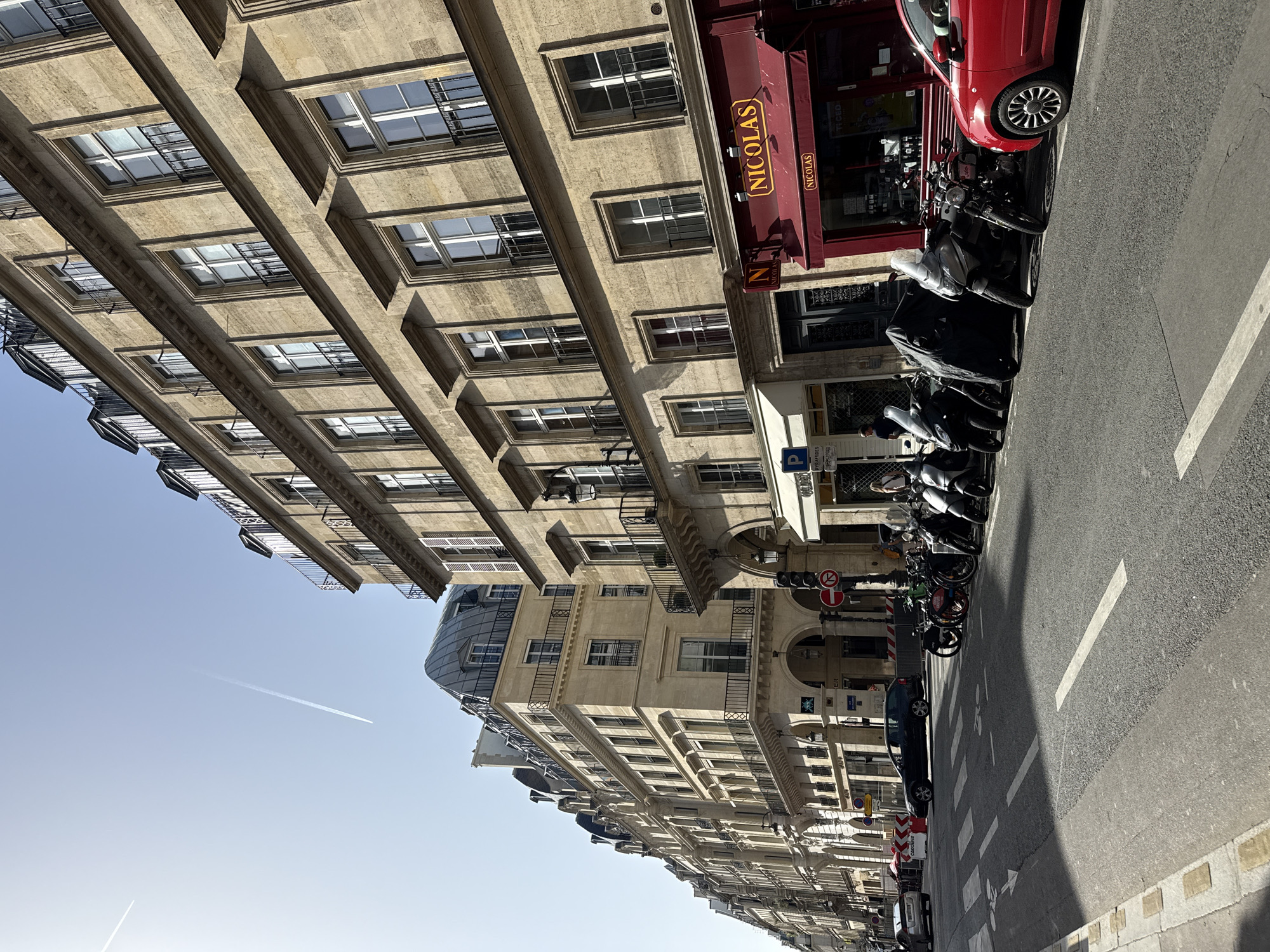 The image depicts a street scene in Paris, France. The view is along a street lined with Haussmann-style buildings. These buildings are characterized by their uniform facade, light-colored stone, and multiple windows. A "Nicolas" wine shop is visible with its red awning. Scooters and a red car are parked along the edge of the sidewalk next to the street. The street itself is asphalt with white lane markings. In the sky, a contrail from an airplane cuts across the blue sky.