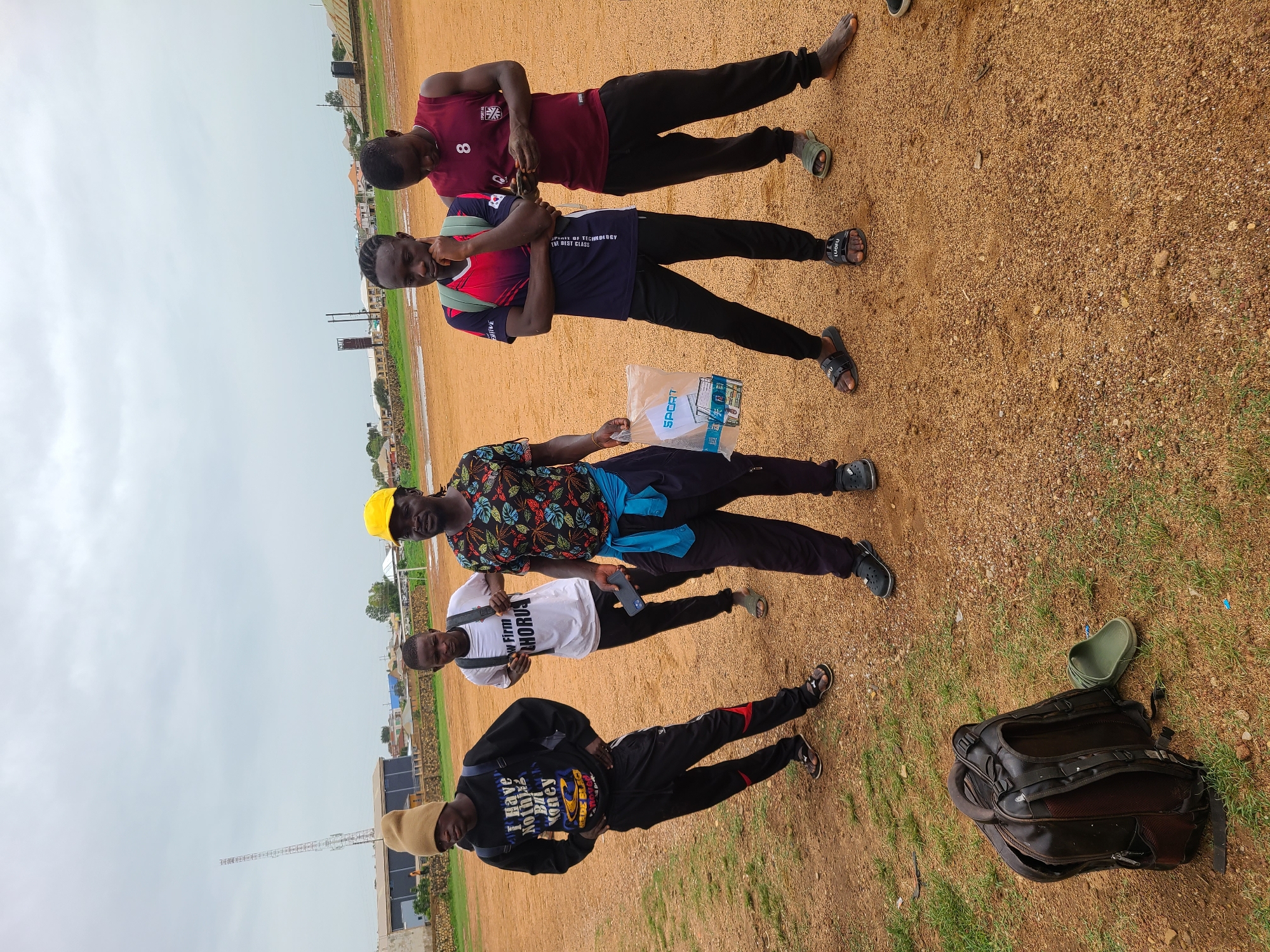The image depicts a group of five young men posing outdoors on a dirt and grass field, likely a sports field. They are arranged in a vertical line across the frame.
The first man, at the top, is wearing a maroon shirt with the number 8 on it and black pants with green shoes. The second man is wearing a red and blue shirt with black pants and black sandals. He has a small backpack on. The third man is wearing a patterned shirt and a yellow hat, with a blue cloth tied around his waist. He holds a clear plastic bag marked with "SPORT." He has on black pants and black shoes. The fourth man wears a white shirt with "New Firm CHORUS" printed on it, black pants, and sandals. The fifth man, at the bottom, wears a black long-sleeved shirt with a graphic design and text that includes "I have nothing to prove but I have Money," black pants with red accents, and a tan hat.
To the right of the fifth man, there is a black bag and a green shoe.
The background includes buildings and a telecommunications tower, indicating an urban or suburban setting. The sky is overcast. The location appears to be in the city of Unknown, Unknown.
