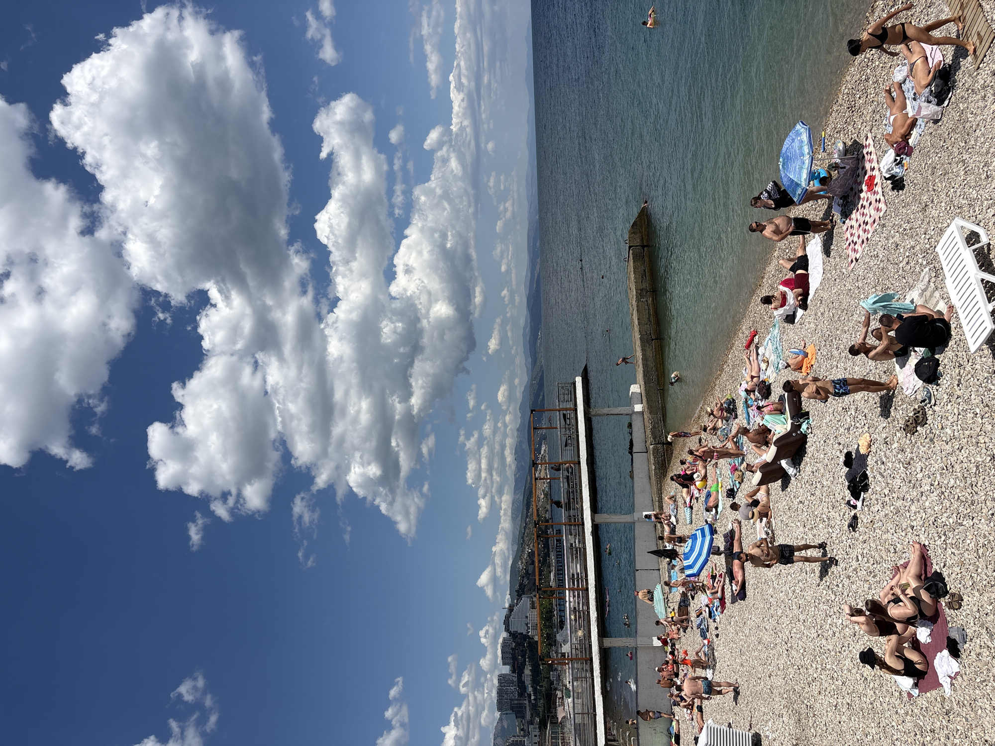 The image depicts a crowded pebble beach in Alushta, Ukraine, on a sunny day with scattered white clouds. Numerous people are sunbathing on towels and lounge chairs. Some individuals are in the water, swimming or floating. A concrete pier extends into the sea, and a raised platform with railings and seating areas is visible above the beach. In the background, the cityscape of Alushta is seen with multi-story buildings and mountains beyond. The sea is calm and blue, meeting a hazy horizon.