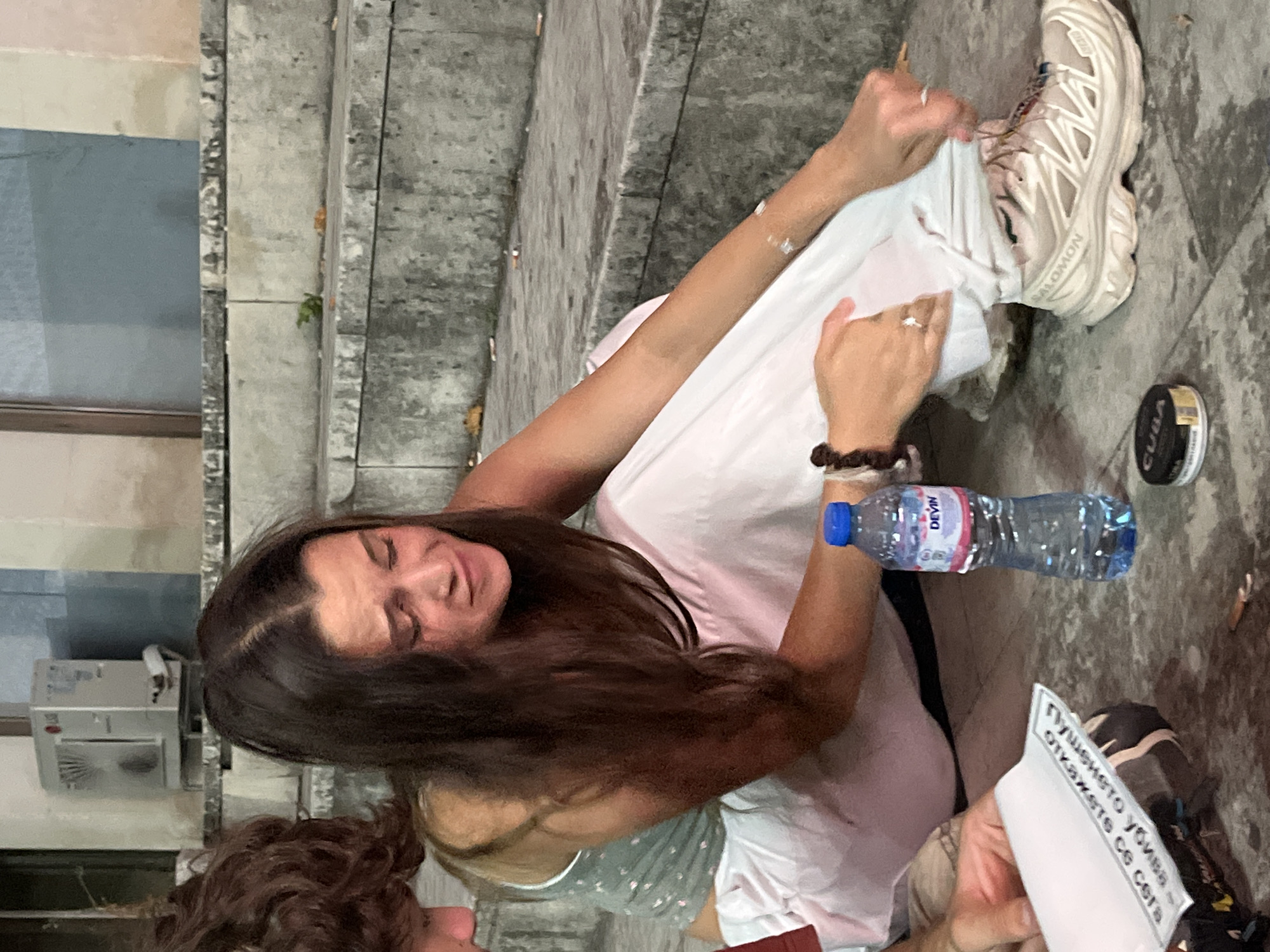 The image depicts a young woman with long, dark hair, crying and sitting on concrete steps. She is wearing a light-colored tank top and white pants. Her right hand is touching her left leg, and her left hand is fiddling with the hem of her pants near her shoe. A bottle of "DEVIN" water and a tin labeled "CUBA" are on the ground near her. In the background, an outdoor air conditioning unit is visible on a ledge. The steps appear worn and stained, and there are cigarette butts scattered on them and the ground. The setting appears to be an outdoor public space, possibly a park or an area with tiered seating. Based on the visible elements, the location cannot be definitively determined as Vratsa, Bulgaria.