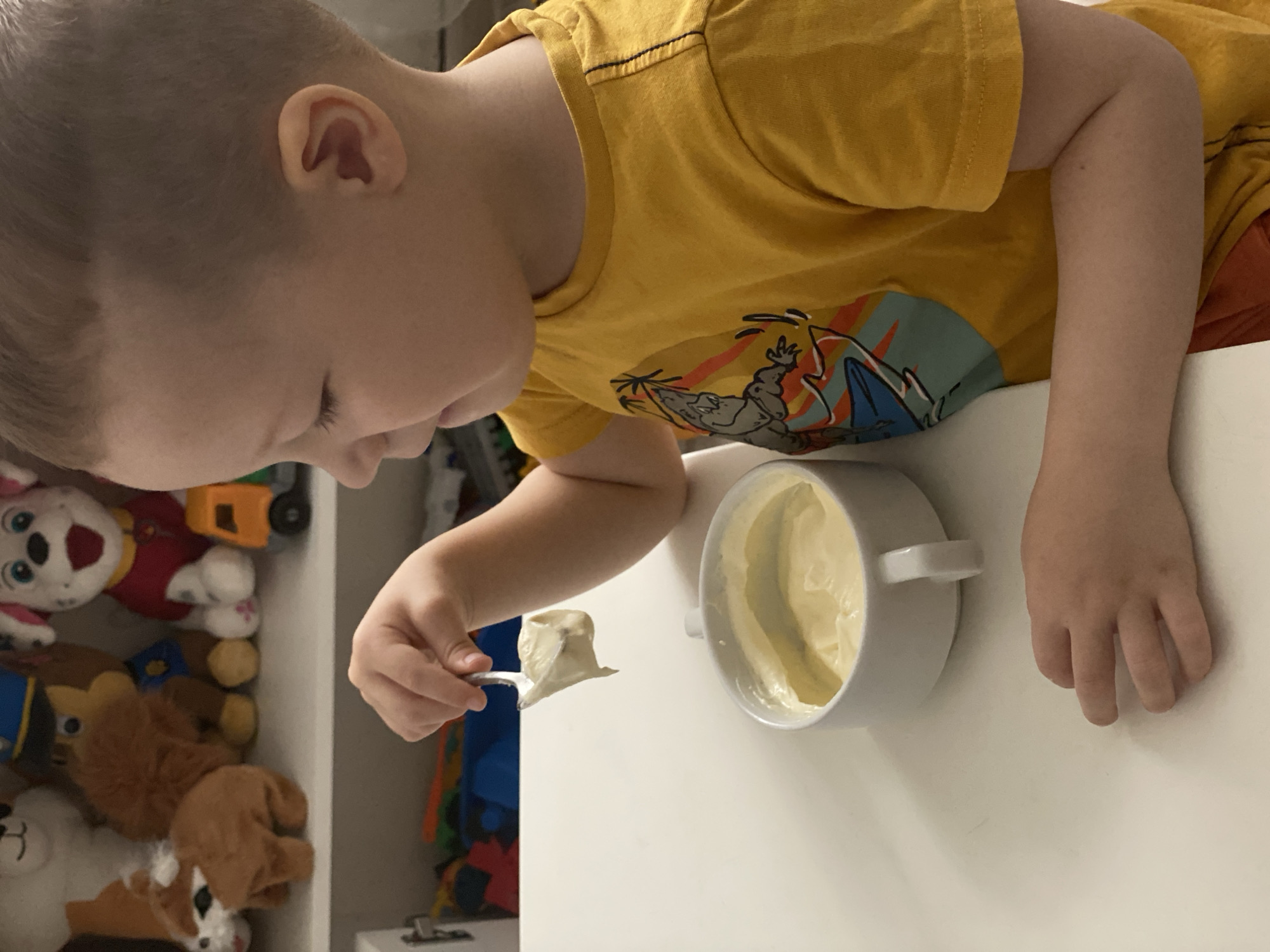 A child wearing a yellow t-shirt with a graphic design is holding a spoonful of what appears to be cream cheese or a similar spread. The child is positioned near a white bowl containing more of the same spread. In the background, there's a shelf filled with stuffed animals. The location in Sochi, Russia, cannot be determined from the visual information provided.