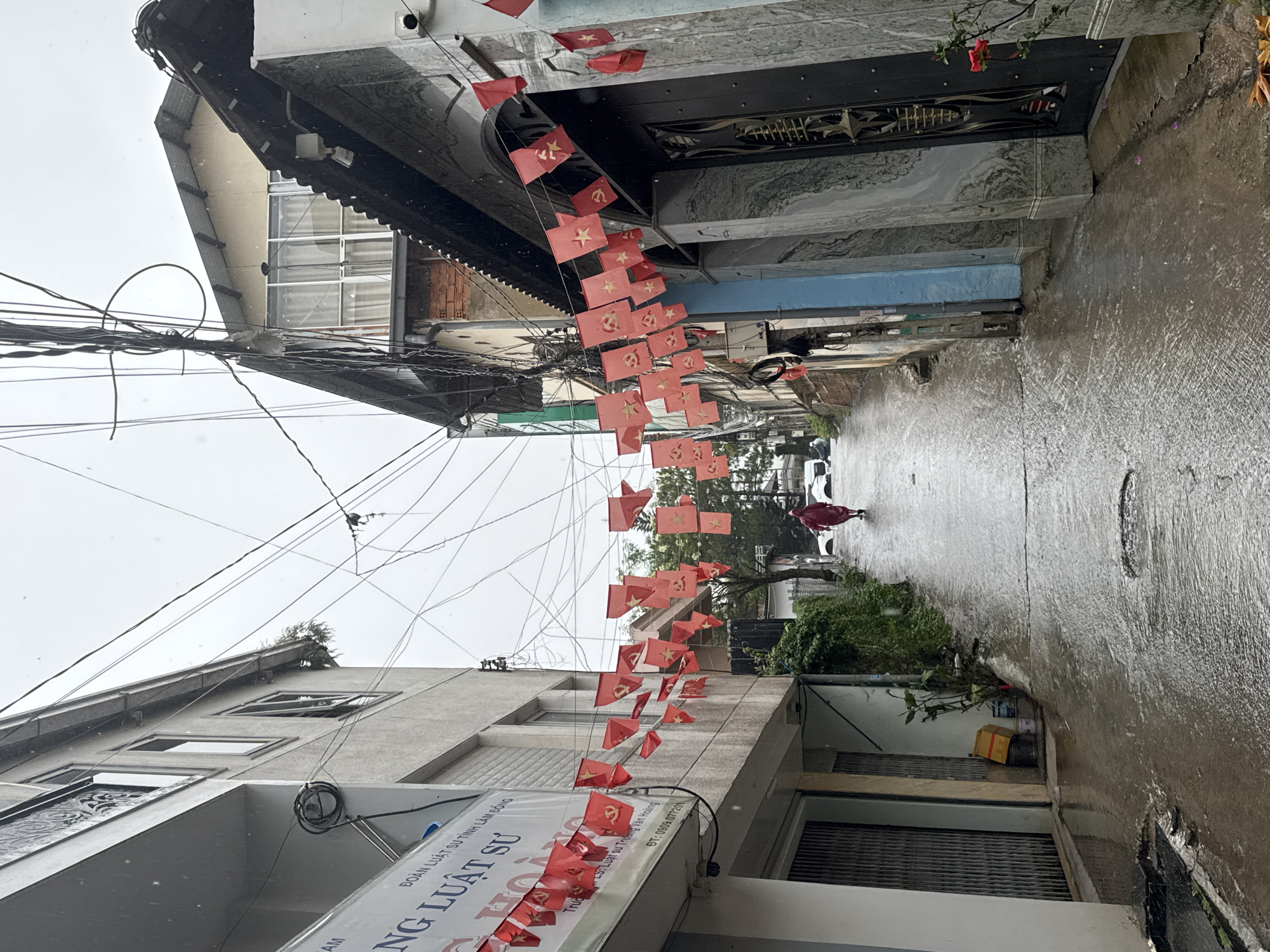 This is an eye-level shot of a street in Da Lat, Vietnam, taken on a rainy day. The street appears narrow and wet, reflecting the overcast sky. The perspective is aligned along the street, with buildings flanking both sides. Above the street, numerous red flags are strung, likely for a celebration or national holiday. The flags feature communist symbols: a yellow star on one variant and a hammer and sickle on another.
The buildings appear to be residential and commercial, with a mix of architectural styles. One building displays signage in Vietnamese, indicating it is a law office: "ĐOÀN LUẬT SƯ TỈNH LÂM ĐỒNG," and lists a phone number. A single individual, wearing a red jacket, is walking down the street, adding a human element to the scene.
The overall atmosphere is somewhat somber due to the weather, but the flags suggest a festive occasion. Power lines are visible overhead, further emphasizing the urban environment.