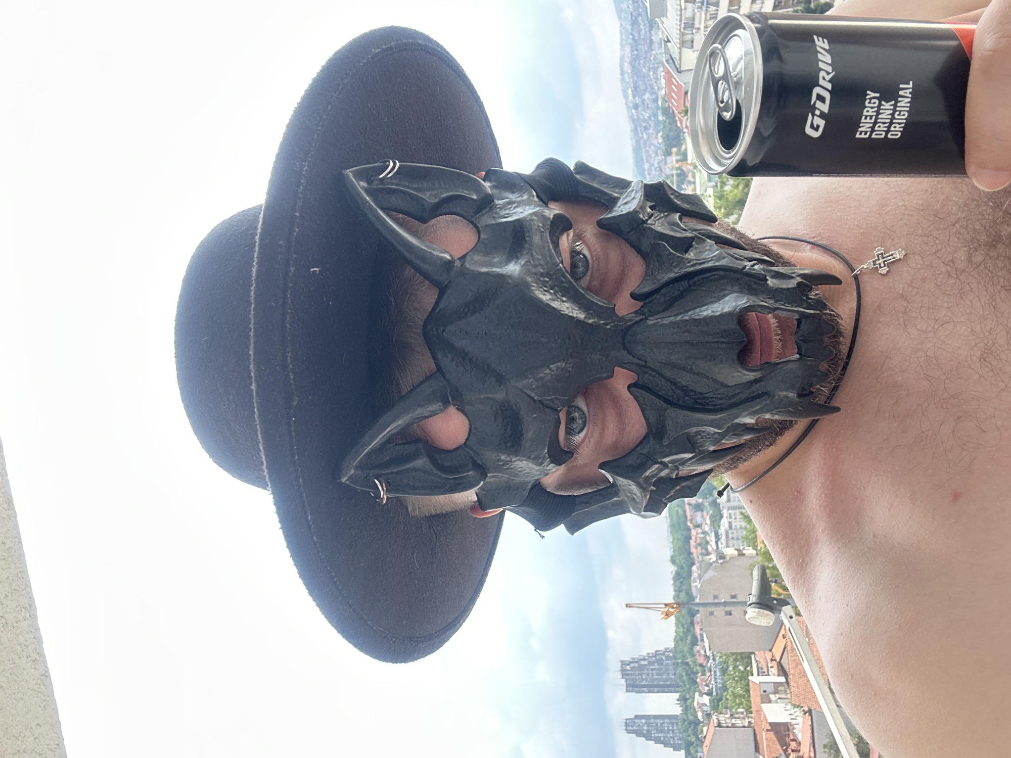 The media file is a close-up, rotated shot of a person wearing a black mask and a black hat. The mask covers the upper half of their face and appears to be made of a rigid material with pointed features, resembling a demon or creature. The person's eyes are visible through the mask's eyeholes and appear to be looking directly at the camera. They are also holding a black can of "G-DRIVE ENERGY DRINK ORIGINAL" in their right hand. A silver cross pendant on a black cord is visible around their neck. The background is out of focus, showing a cityscape with buildings and trees under a cloudy sky, suggesting an urban setting. The lighting suggests it is daytime. The image is taken from a high vantage point, possibly a balcony or rooftop, given the view of the city below. The overall mood is somewhat mysterious or edgy due to the mask.