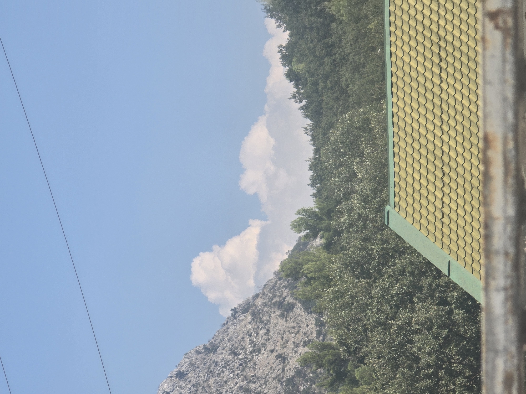 The image depicts a mountainous landscape with a clear blue sky and scattered white clouds. In the foreground, the roof of a building with yellow, wavy tiles is visible. The roof is bordered by a green edge. Behind the roof, a dense forest of green trees stretches across the middle ground. The background features a rocky, grey mountain. Power lines are visible in the upper part of the sky. The scene appears to be an outdoor environment, likely in a location with natural beauty, possibly a suburban or rural area adjacent to mountains, like the outskirts of Podgorica, Montenegro, known for its proximity to mountains such as Lovćen. There are no people or animals in the image. The image is a still photograph captured during daylight.
