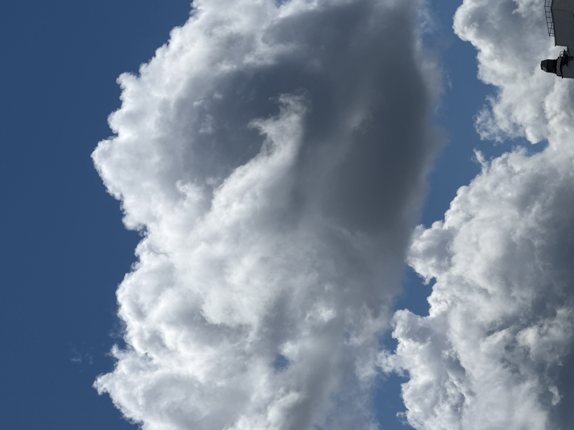 The image captures a dramatic sky filled with a variety of clouds over Yekaterinburg, Russia. The dominant feature is a large, dark, cumulus cloud formation dominating the center of the frame, with lighter, fluffy cumulus clouds filling the lower portion of the image and parts of the upper frame. The sky is a clear, vibrant blue. In the bottom right corner, the very top of a white building with a dark chimney or vent is visible. There are no people or visible actions depicted in the image. The environment is an outdoor scene focused on the atmospheric conditions.