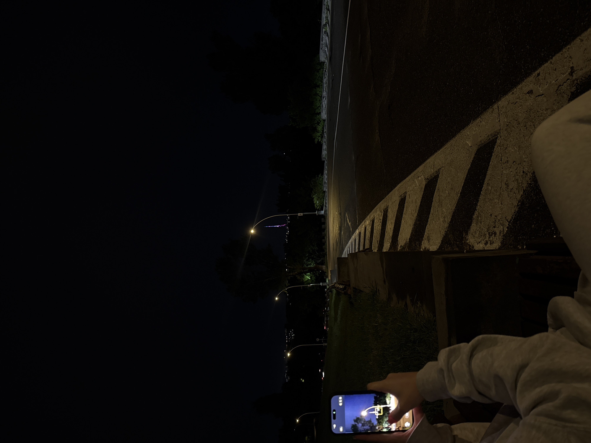 A person wearing a light gray hoodie is holding a smartphone in their right hand. The smartphone's screen displays a daytime image of a street scene, likely taken recently. The person is standing on a dark paved surface next to a concrete barrier with a diagonal white stripe pattern. The sky is very dark, indicating it is nighttime or a very dimly lit location. Street lights are visible in the distance, illuminating the area. Based on the visible landmarks and the style of street lighting, the location could be in Yerevan, Armenia. The person appears to be taking a picture of the night scene with their phone.