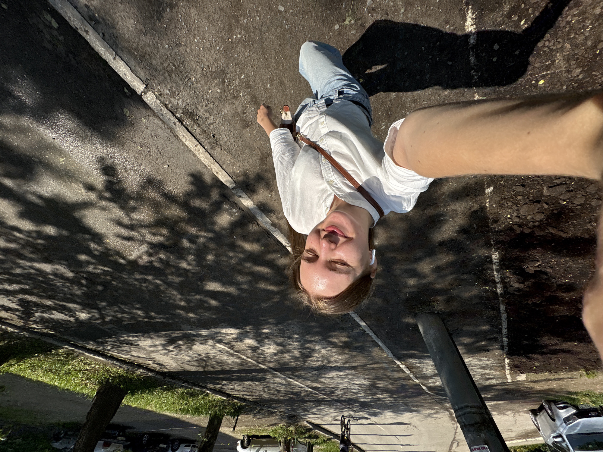 A selfie photograph taken in Perm, Russia shows a young woman standing on a city street. She is wearing light blue jeans, a white button-down shirt, and sandals. A brown leather purse strap crosses her chest. She has light brown hair and wears a white earbud in her left ear. Her eyes are closed and her tongue is sticking out. The camera is held at arm's length, including part of her right arm in the shot. The street is asphalt and shows tree shadows. A curb and sidewalk run along the left side of the image. A utility pole stands to the right. Cars and a bicycle are visible in the background.