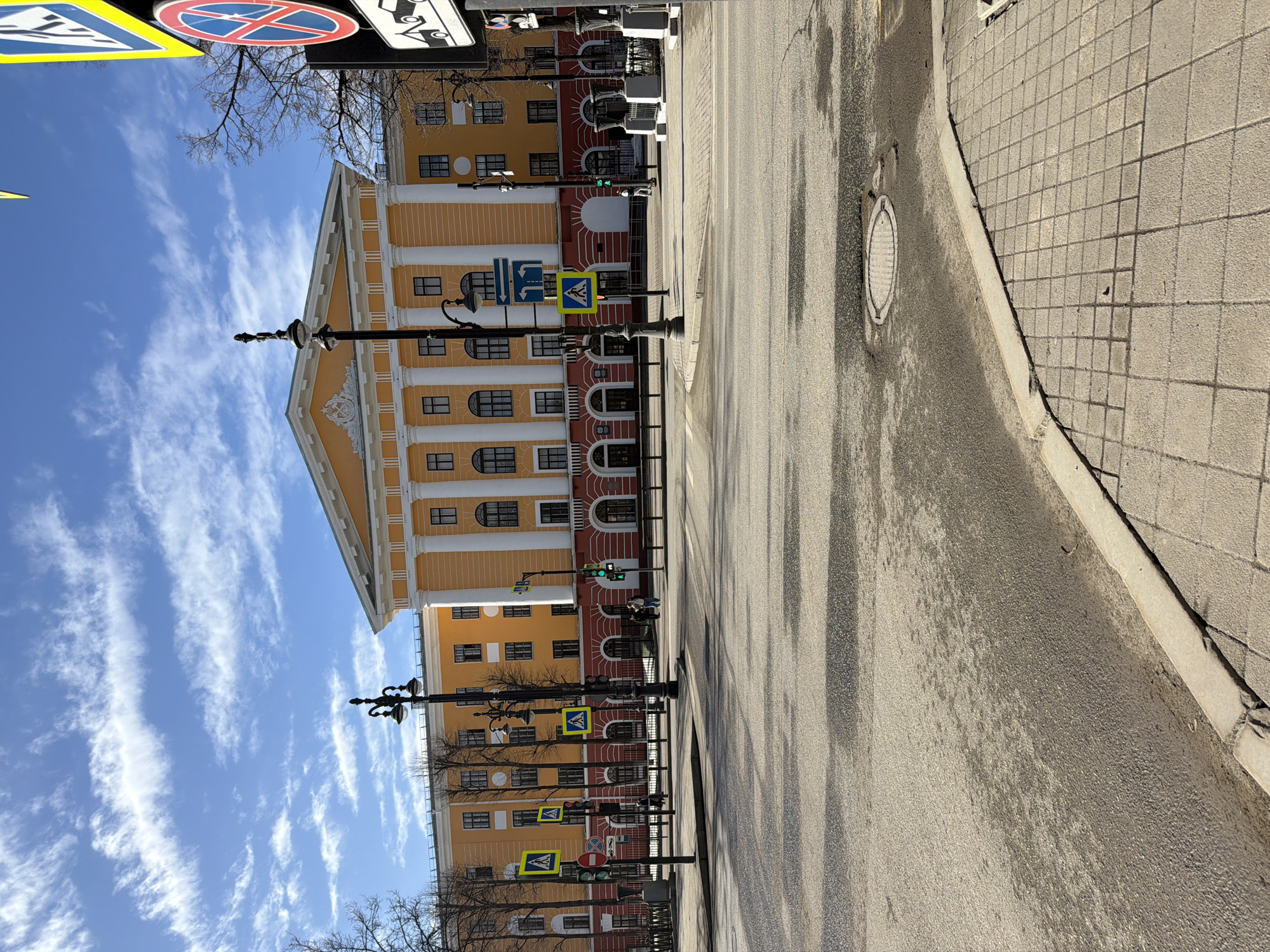 The image depicts a daytime street scene in Perm, Russia, specifically looking towards the Perm Academic Theatre-Theatre at Lenin Street, 53. The prominent subject is a large, multi-story building featuring a pale yellow upper facade with numerous white classical columns and a central pediment containing an emblem. The ground level is painted a deep red, with arched entrances and windows. In the foreground, a wide, asphalt street is largely empty, with traces of dirt and debris. A tiled pedestrian sidewalk and concrete curb occupy the lower right.
Multiple urban elements are present: ornate black lampposts with twin lights, traffic light poles (one displaying a green pedestrian signal), and various road signs. These signs include pedestrian crossing indicators (blue squares with white walking figures), a "No Stopping" sign (red circle with blue background and red diagonal cross), a tow-away warning sign (white rectangle with black tow truck icon), and a blue directional sign indicating options for straight or right turns. Bare deciduous trees line the street in front of the building. Several indistinct individuals are visible walking on the far sidewalks. The sky is clear blue with scattered white, wispy clouds. Shadows on the street surface indicate bright sunlight.