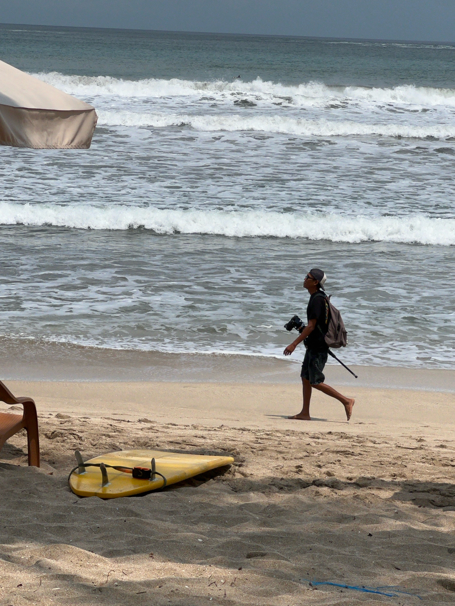 This is a medium shot, taken from a slightly elevated angle, of a beach scene. The foreground shows a yellow surfboard lying flat on the sand, with a dark brown chair to its left. A person is walking along the shoreline from right to left. This individual is wearing a dark t-shirt, denim shorts, a baseball cap, and sunglasses. They are carrying a backpack and a camera with a tripod. The middle ground is occupied by the ocean, with several white waves rolling towards the shore. In the background, the sea extends to the horizon under a cloudy sky. The overall atmosphere is that of a typical day at Kuta Beach, Indonesia, known for its surf and sandy shores.