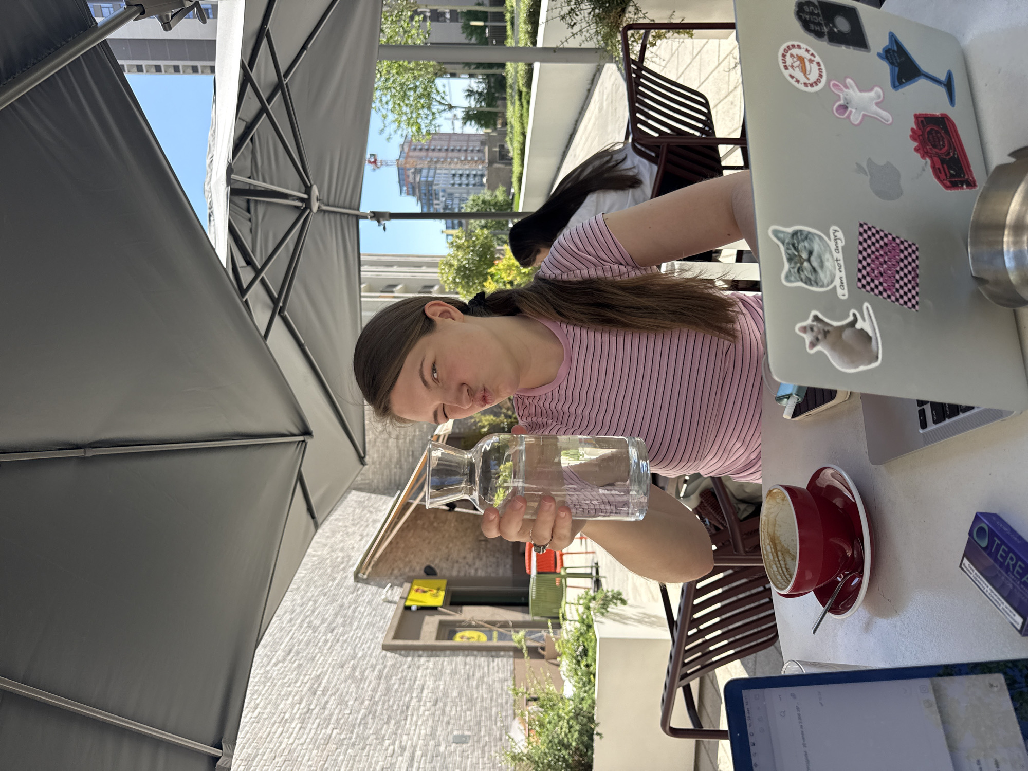 The image depicts a woman sitting at an outdoor table in Belgrade, Serbia, identifiable by the architecture in the background. She is holding a glass water carafe in her hands. She has dark hair pulled back in a ponytail and is wearing a pink and white striped shirt. On the table, there is a laptop with stickers, a red coffee cup and saucer with a spoon, a tablet and a pack of cigarettes. An umbrella shades the table, and there are buildings in the background. Another person is sitting at a table behind the woman.