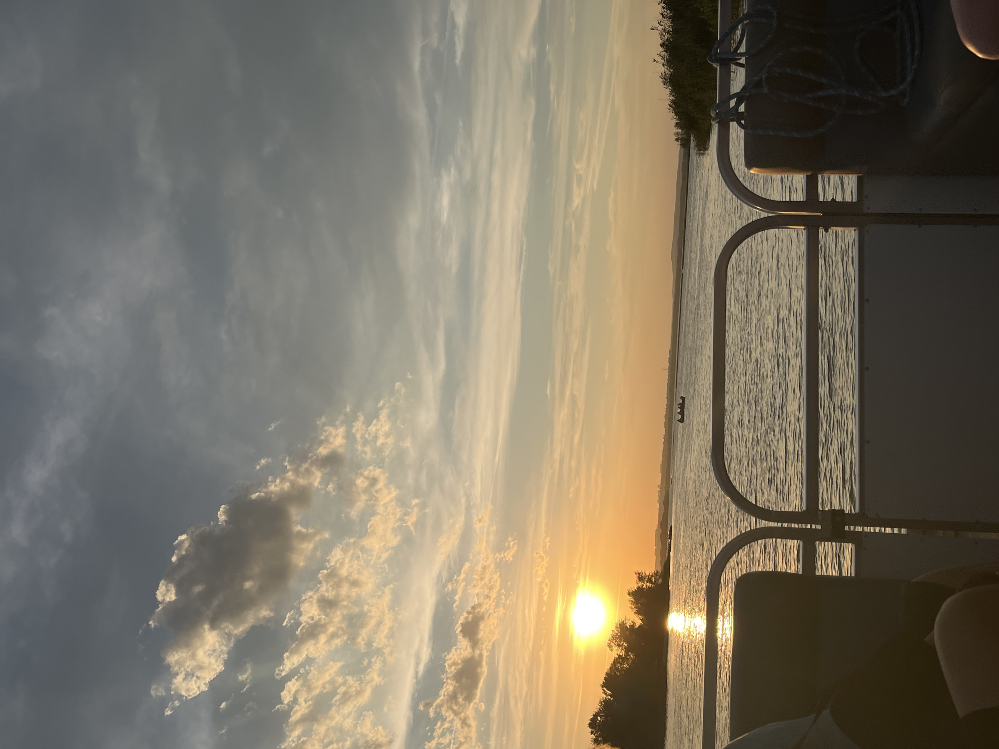 The image depicts a serene sunset over a body of water, viewed from the deck of a boat. The sun, a bright orb casting a warm glow, is low on the horizon, partially obscured by a silhouette of trees on the left side of the frame. The water reflects the golden hues of the sunset, with gentle ripples disturbing its surface. In the distance, several small boats with people can be seen. The sky is a gradient of soft blues and oranges, with scattered clouds catching the light. The foreground of the image shows parts of the boat's interior, including seating and railings, suggesting a leisurely boat trip. There are no discernible landmarks that would indicate the specific location of Shumeika, Russia.