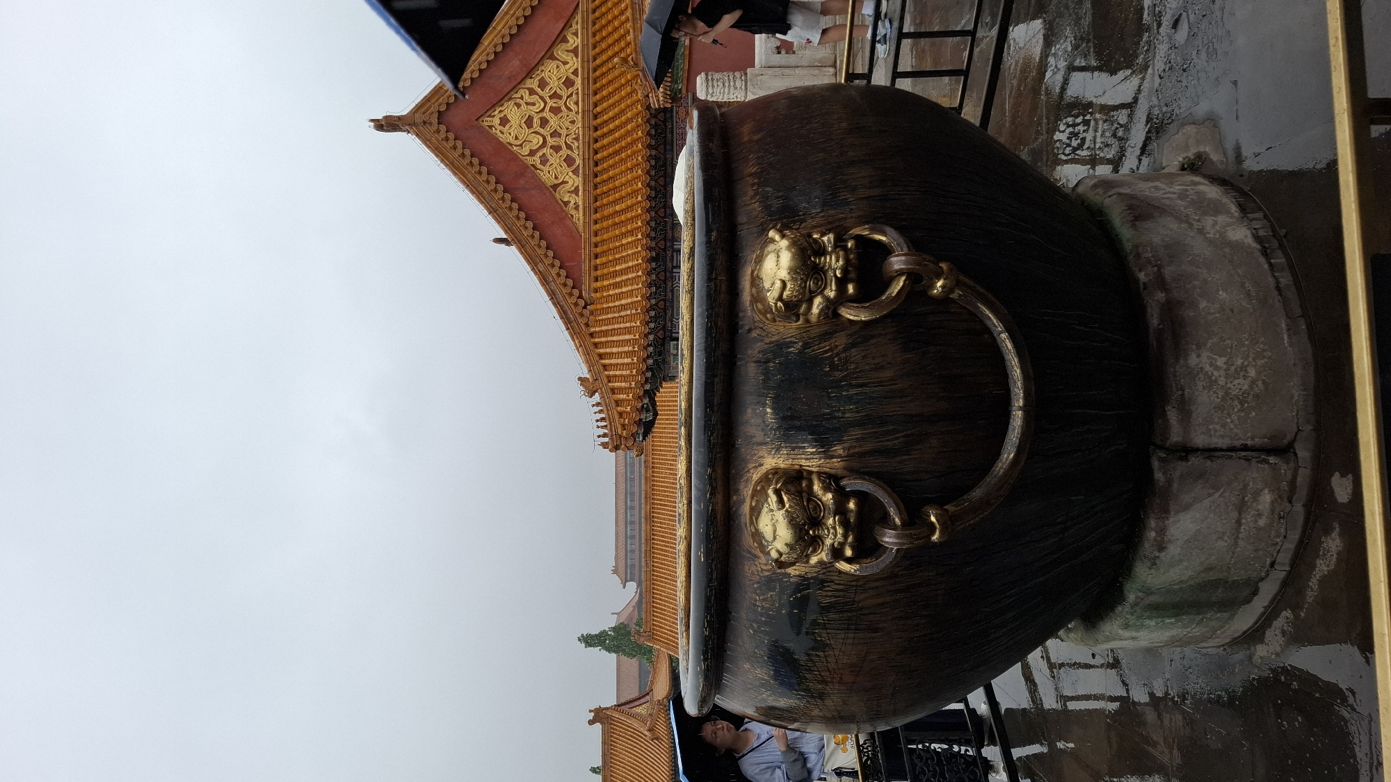 The image depicts a large bronze cauldron with ornate lion head handles, likely located in the Forbidden City in Dongcheng District, China. The cauldron rests on a weathered stone base. Behind it, the traditional yellow-tiled rooftops of the palace buildings rise against a cloudy sky. A person holding an umbrella is visible beneath the cauldron.