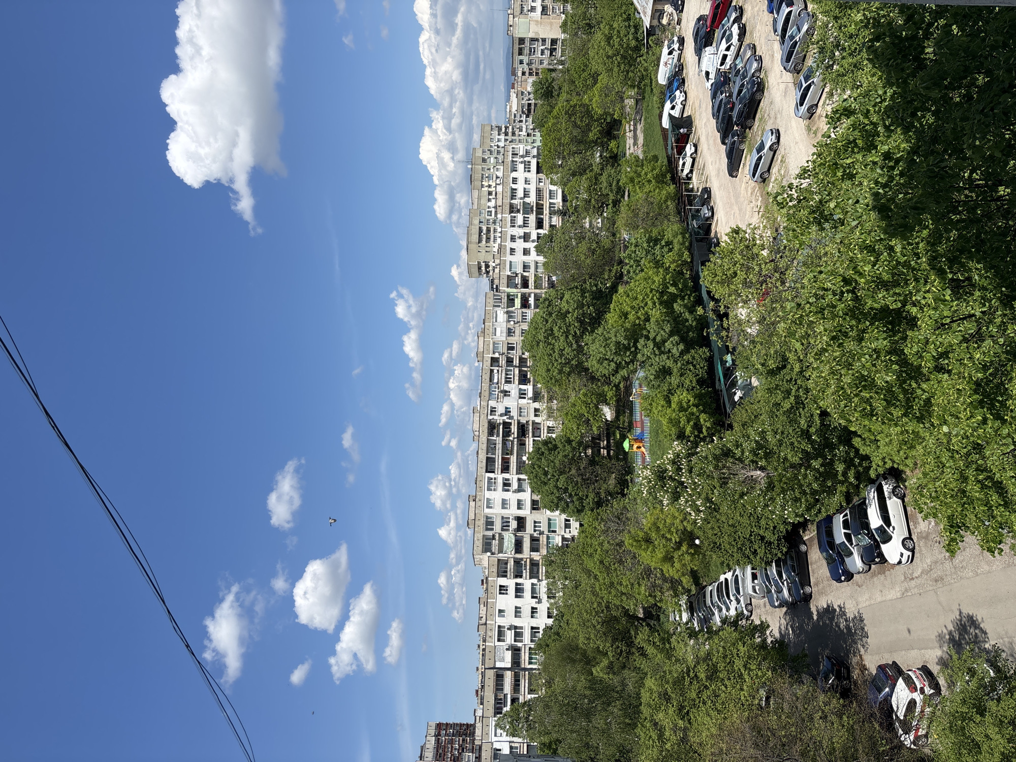 A street scene in Sofia, Bulgaria, viewed from an elevated position. Tall apartment buildings dominate the skyline, contrasted by a vibrant blue sky with scattered clouds. A parking area is filled with cars, interspersed with lush green trees. A playground with colorful equipment is visible. Power lines are also present in the scene.