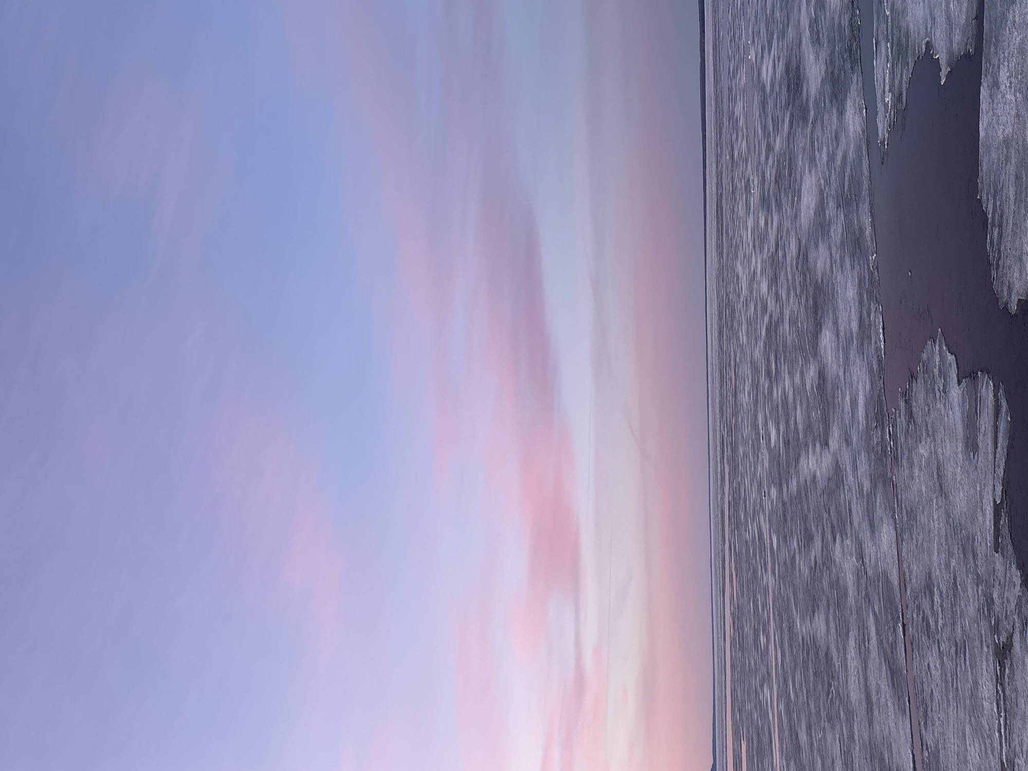 The image presents a tranquil winter landscape captured at what appears to be dawn or dusk in Chaikovsky, Russia. The scene is horizontally divided between a vast, colorful sky and a partially frozen body of water.
The upper portion of the image is dominated by the sky, displaying a soft and expansive palette of pastel hues. Pale blues blend seamlessly into delicate pinks and purples, indicating either the first light of sunrise or the fading light of sunset. Wispy, streaky clouds traverse the sky, especially towards the right, adding to the ethereal and diffused lighting.
Below the horizon, a large body of water is visible, covered in various forms of ice. The ice sheet extends across the frame, appearing textured and slightly irregular, with patterns suggesting refrozen surfaces or subtle ridges. Towards the right side of the image, a distinct area of open, dark water breaks the ice expanse, featuring an uneven, jagged edge where the ice has either melted or broken away. A faint, distant shoreline or landmass might be discernible as a thin dark line on the far left horizon.
No people, specific activities, or man-made objects are visible in the serene setting. The overall atmosphere conveys peace and the cold beauty of a natural environment during a transitional light period. There is no visible text in the image.