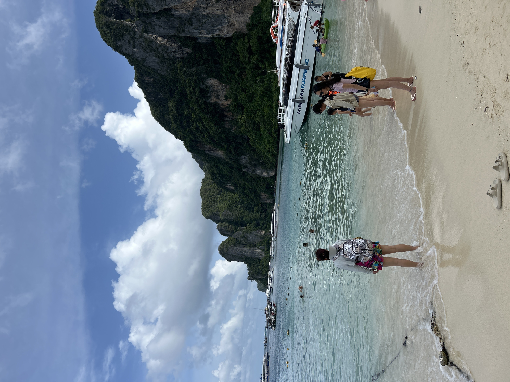 This is a photograph of the beach and ocean at Ao Nang, Thailand. The image is taken from a low angle, with the camera focused on the shoreline and the water. In the foreground, there is a sandy beach with a person standing in the shallow water, facing away from the camera. The person is wearing a backpack and a colorful skirt. Several other people are visible in the water and on the beach in the midground, including a group of young women standing on the sand and a few people swimming or wading.
The water is clear and turquoise, with small waves lapping at the shore. Several boats are anchored in the bay, including speedboats and a larger tour boat with the text "ANDAMAN SURPRISE" visible on its side. In the background, dramatic limestone cliffs covered in lush green vegetation rise from the sea, characteristic of the Krabi province's coastline. The sky above is a bright blue with scattered white clouds. The scene depicts a typical tourist day at a tropical beach destination.