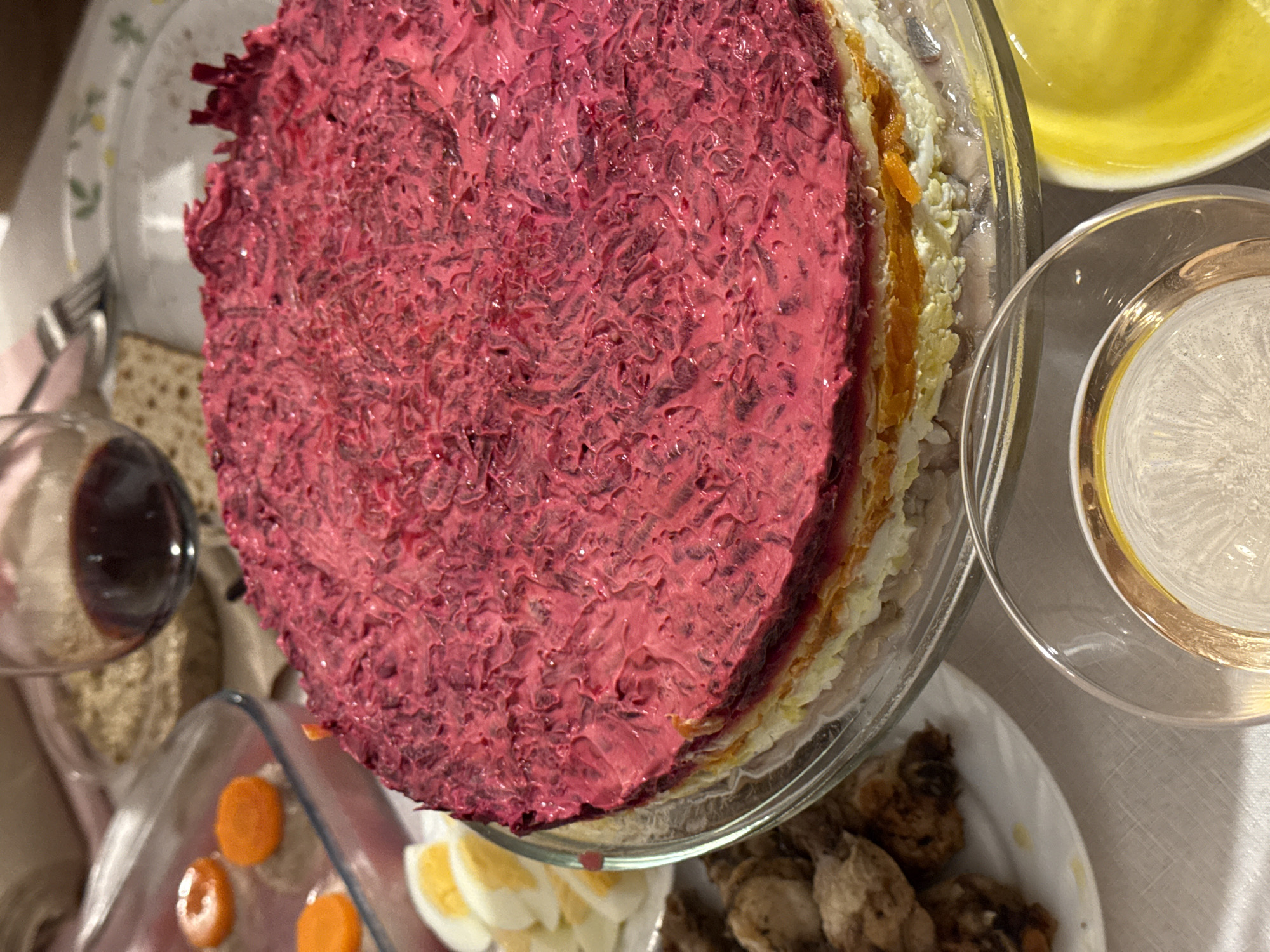 The image depicts a culinary scene, likely indoors on a table setting. The primary focus is a layered salad in a clear glass bowl. The salad is topped with a vibrant pink layer. Additional layers visible include orange, white, and other variations in color, suggesting different ingredients.
Other items on the table include sliced carrots and hard-boiled eggs in a dish, pieces of fish on a plate, a glass of dark liquid, and matzah. There's also a bowl with a yellow liquid, possibly oil or dressing, and a clear glass with a lighter colored liquid inside.
Given the presence of matzah, the scene is likely related to a Jewish holiday meal.
Without additional information, specifying an exact location in Tel-Aviv, Israel, is impossible. However, considering the culinary elements, it is likely a residential dining area within the city. No people are visible in the image.