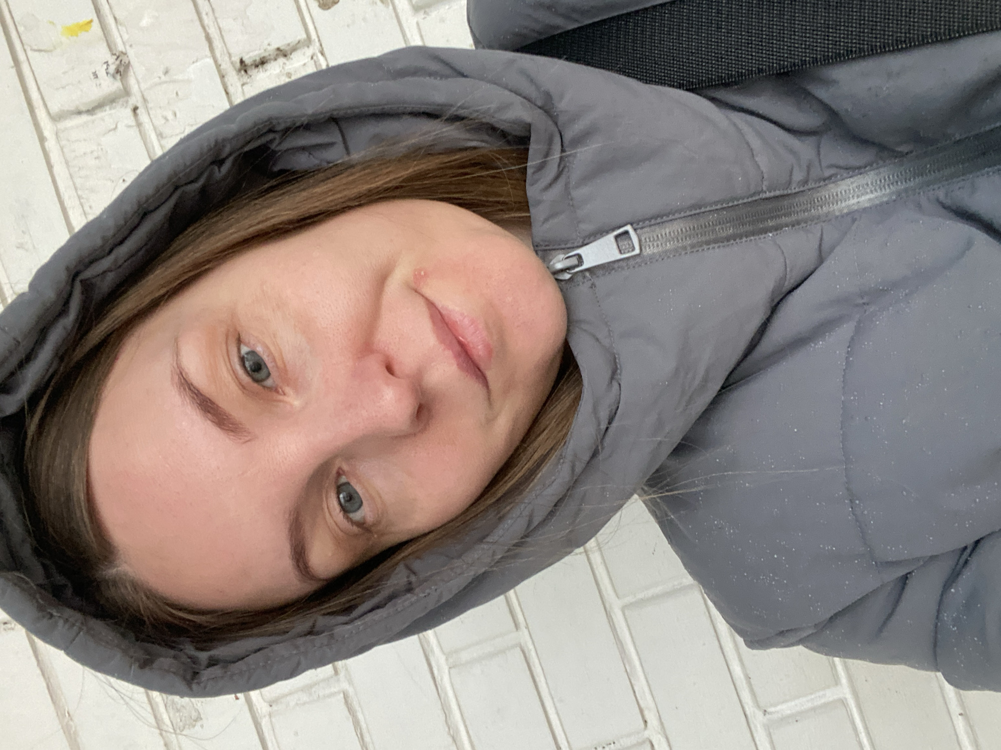 A woman is seen indoors against a wall of white bricks, wearing a gray, padded coat with the hood up. The coat is zipped to the top. The woman is fair-skinned with light brown hair and blue eyes. There is a small blemish on her right cheek. Part of a black strap is visible at the top of the image. The background consists of a white brick wall.
Since the location is Noviy, Russia, this may also be written as Новый, Россия.