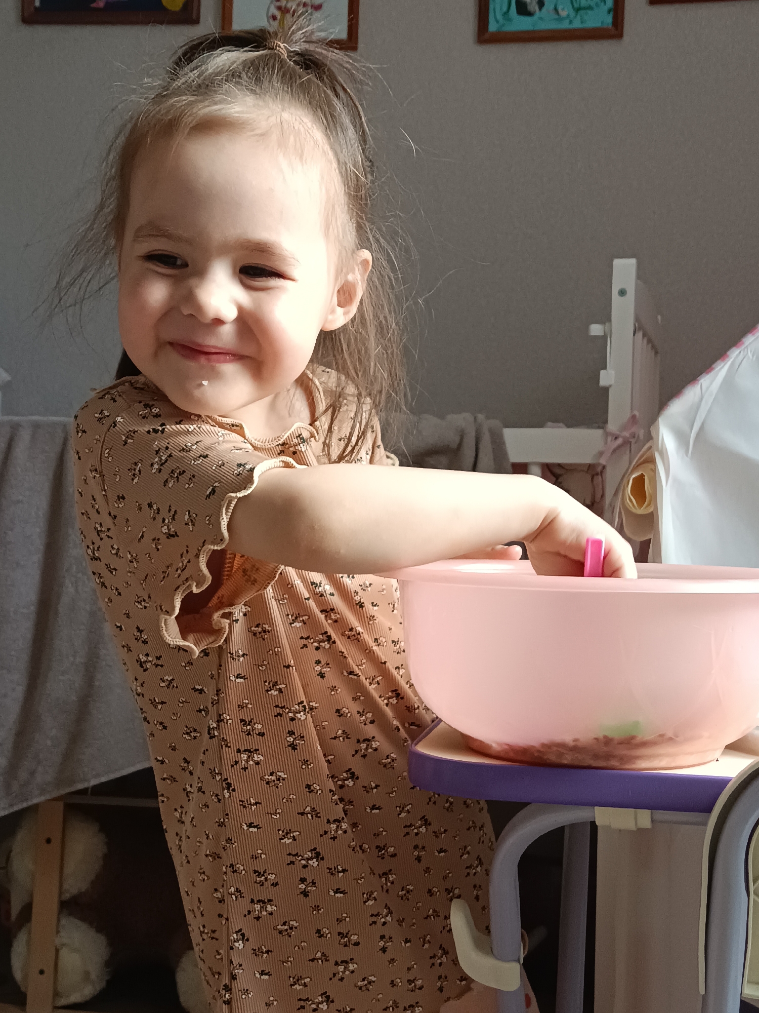 A young child with light brown hair tied in a ponytail is captured indoors, likely in a child's room or play area. The child is smiling broadly, looking towards the left with a joyful expression, and has a small white speck, possibly food, on her chin. She is wearing a light brown or tan long-sleeved top with a subtle floral or patterned texture, featuring ruffled cuffs.
The child's right arm is extended, and her hand is reaching into a large pink plastic bowl, which is placed on a purple and white stand or high chair tray. A pink utensil, possibly a spoon, is visible within the bowl.
In the background to the left, a grey blanket or textile is draped. To the right, part of a white crib or bed frame is visible, along with what appears to be a white fabric structure, possibly a tent or play canopy. Above the child, two framed pictures are hanging on a light-colored wall; one has a blue/green image within a brown frame, and another brown frame is partially visible to its right. At the bottom left, partially obscured by the child's dress, some stuffed animals or toys can be seen.
The scene is brightly lit by natural light, suggesting it is daytime. The child's cheerful smile conveys happiness and engagement in her activity. No discernible text is visible in the image. While the location context is Noviy, Russia, there are no specific visual cues within the image itself to confirm this.