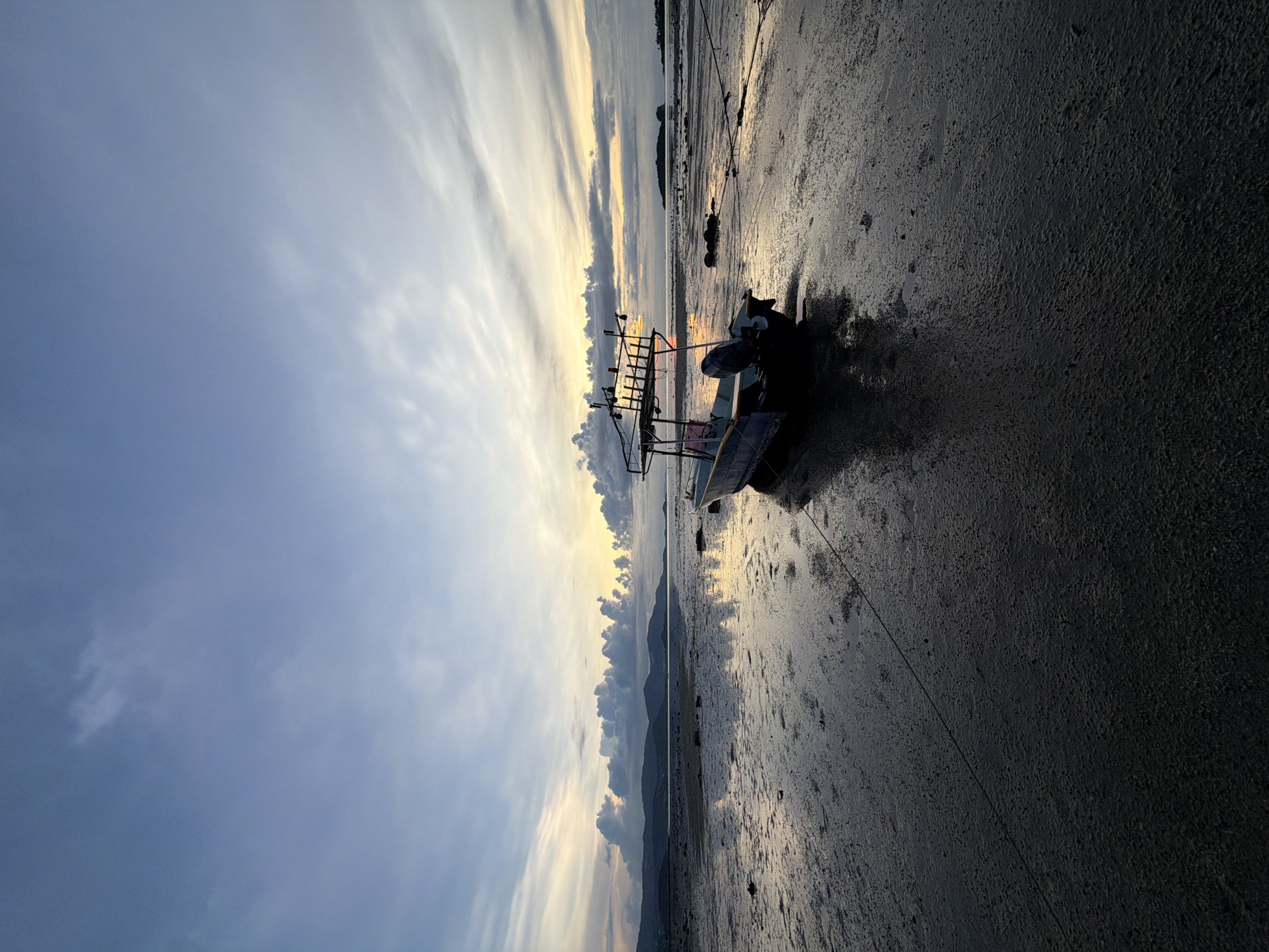 The image depicts a serene coastal scene at Baan Plai Laem, Thailand. The primary subject is a boat resting on a sandy, wet shore during low tide. The boat appears to be a small fishing vessel, with an external motor and a metal frame structure possibly for equipment or storage. It is tethered by ropes to the shore.
The setting is a beach, characterized by wet sand that reflects the sky and the distant landscape. In the background, a line of mountains or hills is visible, softened by the atmospheric haze. The sky is a prominent feature, displaying a mix of soft, elongated clouds illuminated by the warm glow of either sunrise or sunset. This interplay of light and shadow creates a peaceful and somewhat melancholic mood.
The time of day is clearly either early morning or late evening, indicated by the low angle of the sun and the diffused light. The weather appears calm, with no signs of strong winds or rough seas. The location context is explicitly stated as Baan Plai Laem, Thailand, which suggests a tropical coastal environment.
There are no people visible in the frame, emphasizing the solitude of the scene. The overall impression is one of tranquility and stillness, capturing a moment of repose for the boat and the natural surroundings. The wet sand and the gentle hues of the sky are notable details that contribute to the calm atmosphere. There is no visible text in the image.