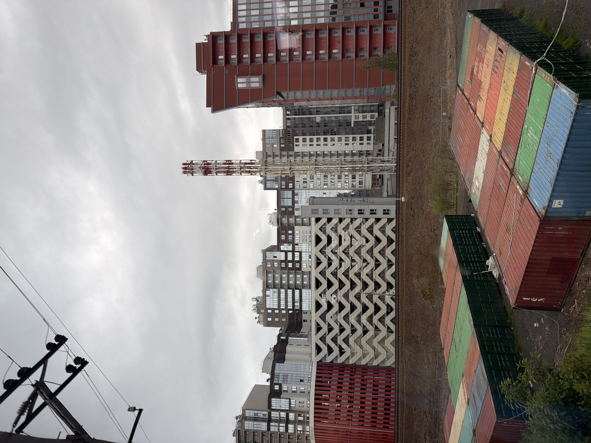 The image depicts an urban landscape under a cloudy sky. In the foreground, several shipping containers are arranged in rows, some are green, others are a mix of red, blue, yellow, and white. A few sparse green bushes are visible to the left. A railway track runs horizontally across the middle ground. Behind the tracks, a series of modern multi-story buildings are visible. These buildings vary in height and design, with some featuring balconies and large windows. A distinctive red building with a geometric facade is on the right, and a tall, white industrial chimney with red accents stands out among the buildings. The overall impression is one of an industrial or semi-industrial area on the outskirts of a city.