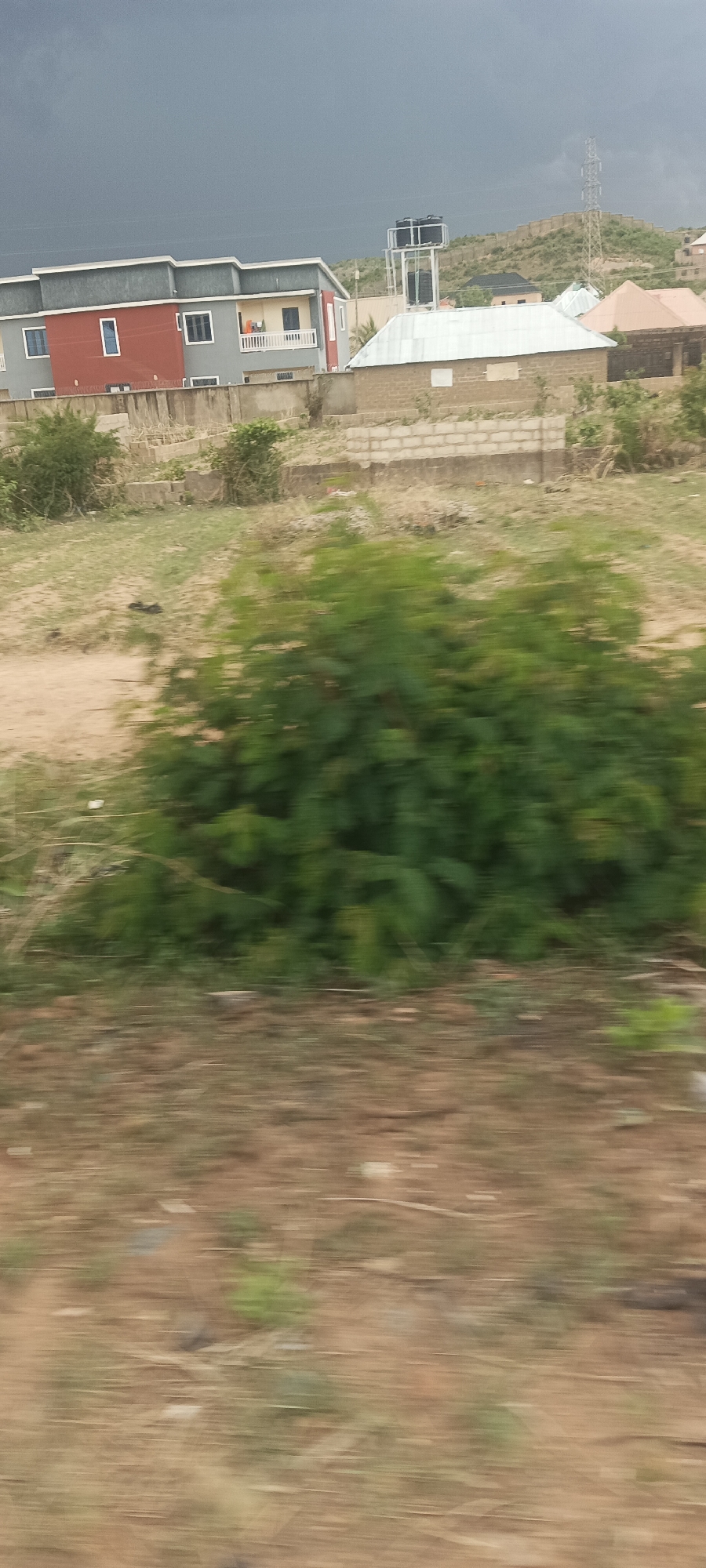 The image depicts a blurred landscape along Rukuba Road, Nigeria, under a dark, overcast sky. In the background, there are several residential buildings, including a two-story house with a red facade and a grey upper level. Adjacent to it is a smaller building with a white roof and a water tower on a nearby hill. A hill with vegetation and a tall electrical transmission tower can be seen in the distance. In the foreground, there is a blurred view of vegetation and a dirt-covered ground, indicating the image was taken while in motion. The overall scene conveys a sense of movement and a slightly gloomy atmosphere due to the dark sky.