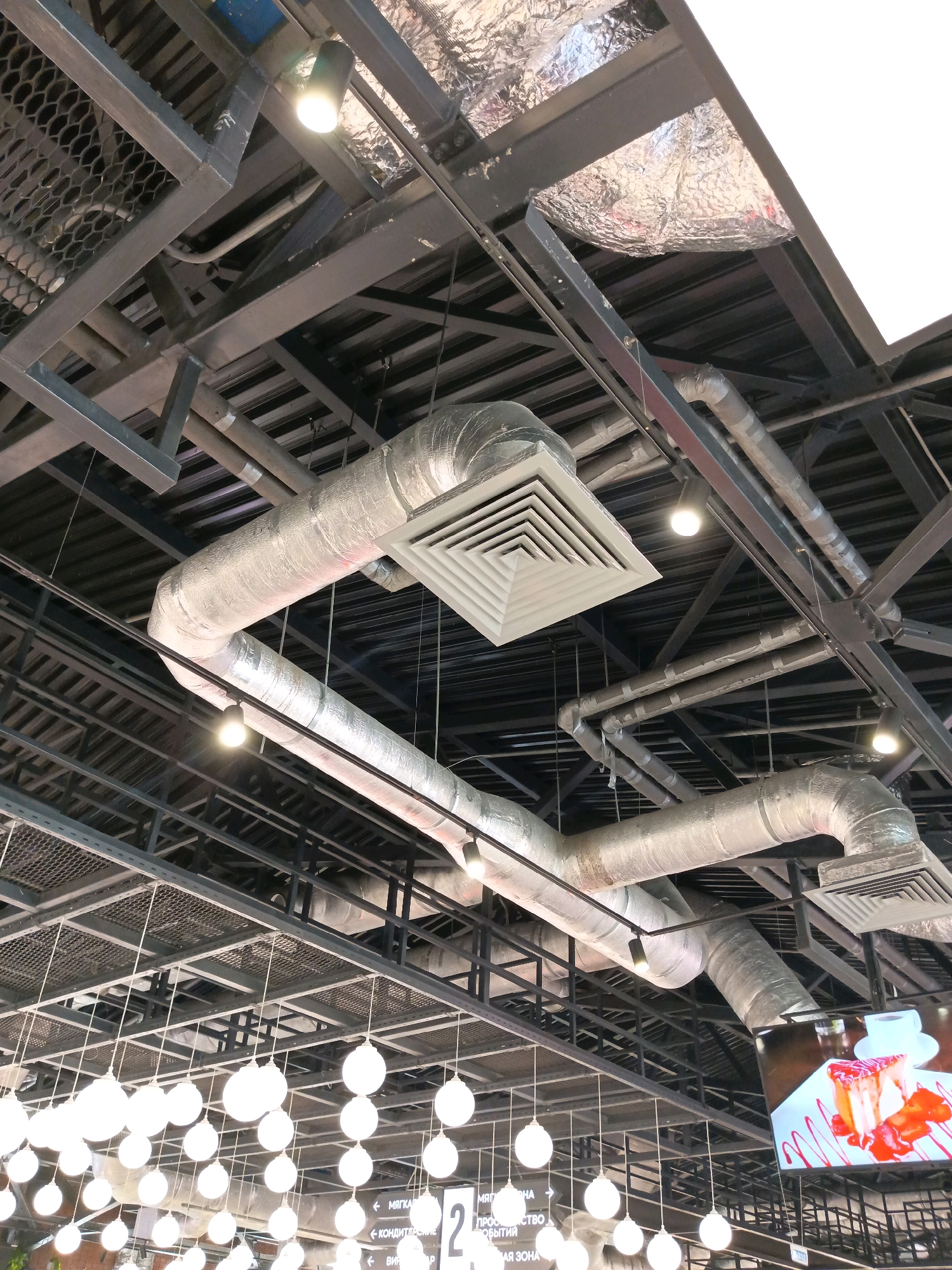 The image shows the ceiling of a commercial space, likely a supermarket or a large store, with exposed ductwork and structural beams. There are several prominent silver-insulated ventilation ducts running across the ceiling, connected to a large square air vent. Rows of spherical white pendant lights hang down from the ceiling. The background shows a complex network of dark metal beams and supporting structures, with some areas of honeycomb-patterned mesh and reflective insulation. A digital screen displaying an image of a slice of cheesecake with strawberries is visible in the lower right corner. There are no people or animals visible in the frame. The overall impression is one of an industrial-style interior design.