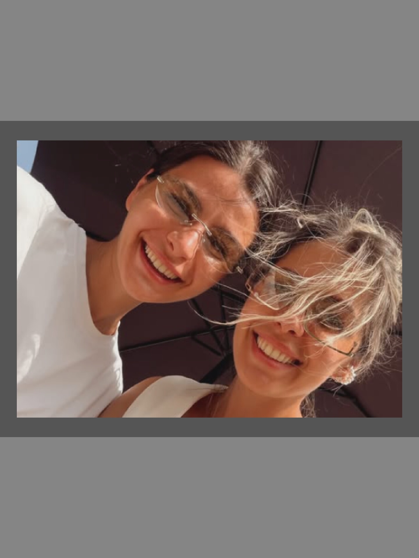 The image shows a close-up, low-angle, overhead shot of two young women smiling at the camera. They are outdoors, possibly under a large umbrella as indicated by the dark, curved background. Both women are wearing sunglasses and are in focus. The woman on the left is wearing a white t-shirt and has dark hair pulled back. The woman on the right has lighter, windblown hair and is wearing a light-colored top and a distinctive white earring. The lighting suggests it is daytime.