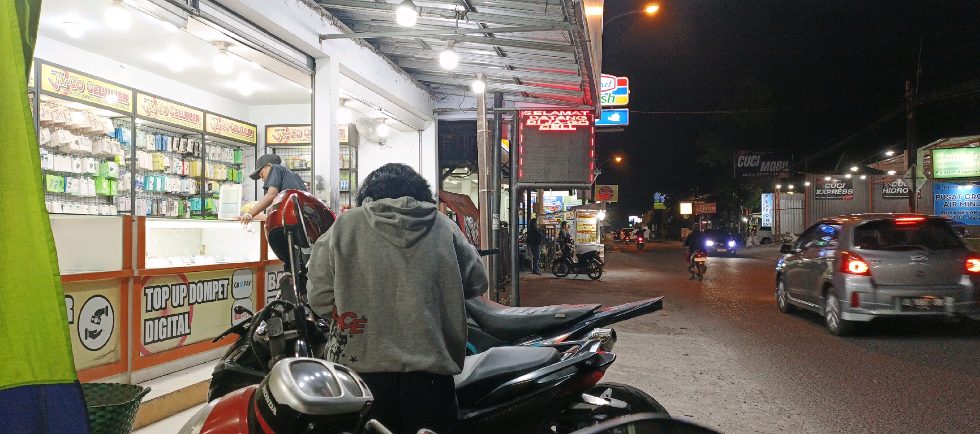 The image captures a nighttime street scene in Sidokarto, Indonesia, likely in front of a mobile phone accessories shop called "JA-GO CELLULER." A person wearing a grey hooded sweatshirt is standing in the foreground, facing away from the camera, with a motorcycle partially visible in front of them. Behind this person, a shop displays numerous phone cases and accessories on shelves. To the right of the shop, a street is visible with several vehicles, including a silver car and motorcycles, in motion. The street is lined with various businesses with illuminated signs, including "SELAMAT DATANG DI JA-GO CELLULER," "CUCI MOBIL," "CUCI EXPRESS," and "CUCI HIDRO." The overall atmosphere is that of a busy commercial area at night, with illuminated storefronts and traffic on the road.