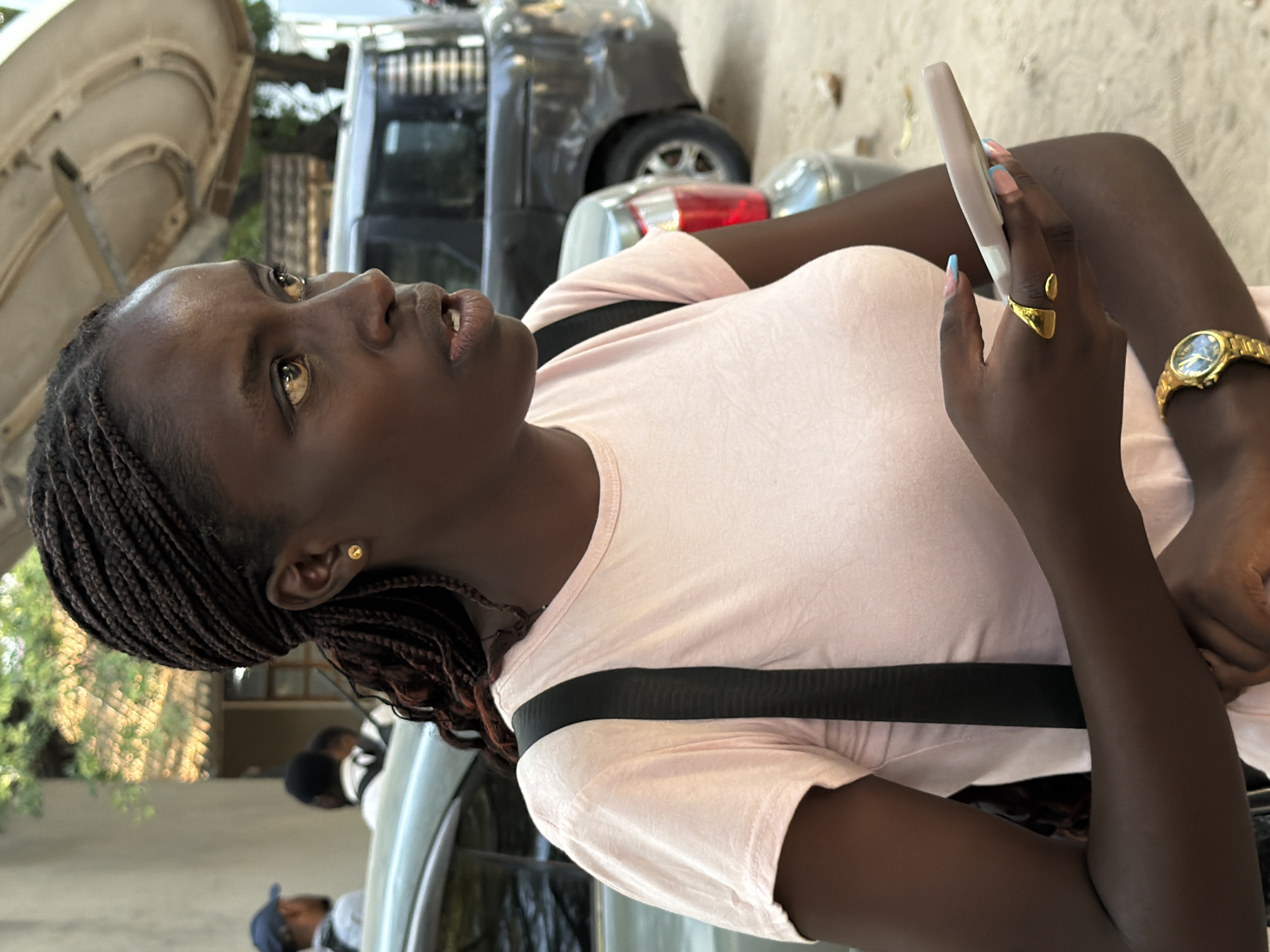 In Dar es Salaam, Tanzania, a young Black woman with braided hair and a light pink t-shirt is holding a light pink smartphone. She is wearing a gold stud earring and a gold ring on her left hand, as well as a gold wristwatch on her right wrist. Her fingernails are painted light blue with white tips. In the background, there are two vehicles, one gray and one dark colored, and a building with trees and foliage. The ground appears to be sandy or dusty.