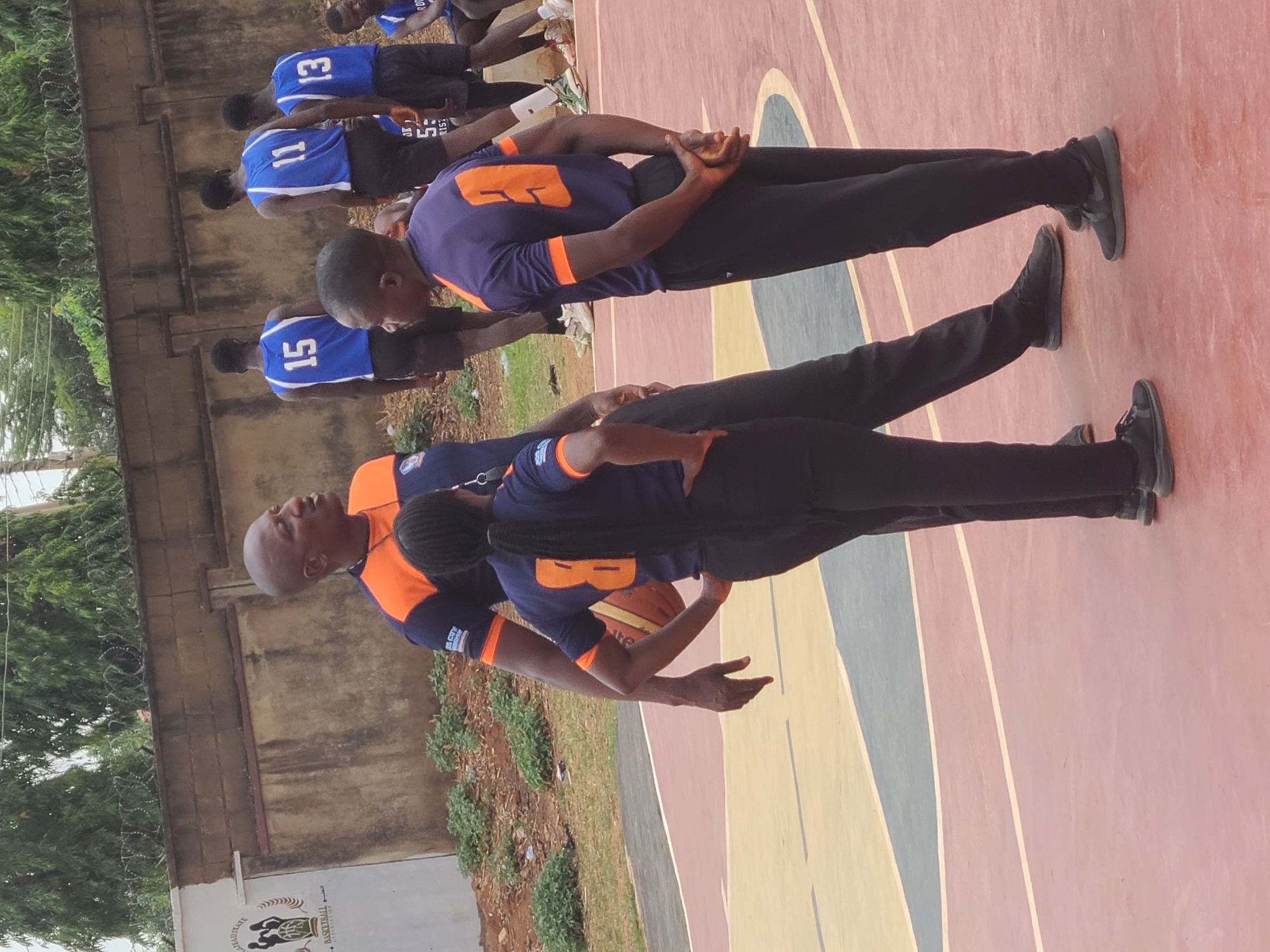 A row of individuals is captured on a basketball court, the location identified by an inscription on a nearby building indicating "Jos City Basketball Association", thus situating the scene in Jos, Nigeria. The individuals are uniformly dressed in blue and orange basketball jerseys. Some are stretching or warming up, their hands clasped behind their backs. The court’s surface is painted with a pattern of beige, grey, and red stripes. In the background, there is a wall topped with barbed wire and some greenery.